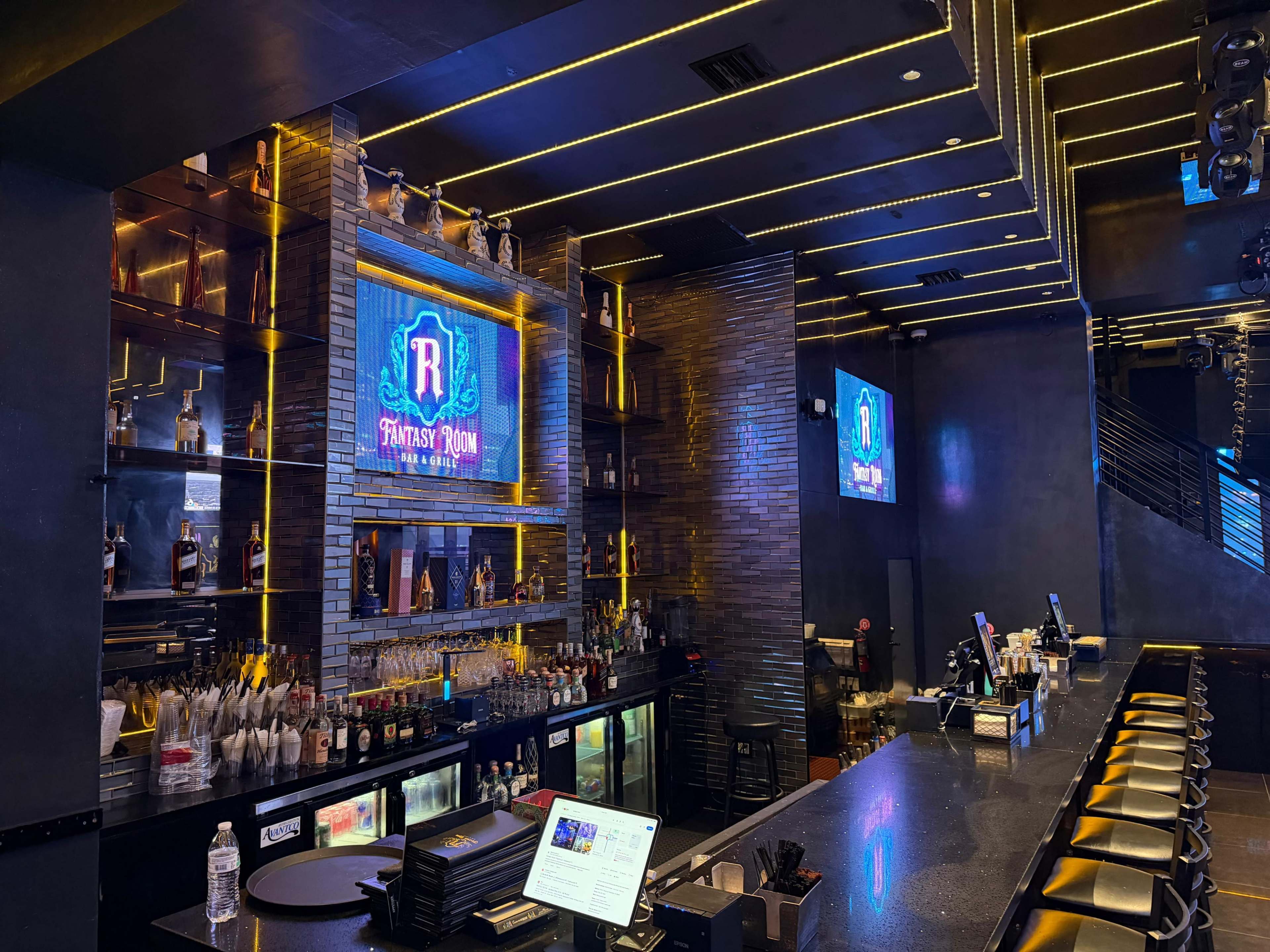 The image shows a modern bar interior with a sleek countertop, illuminated shelves displaying various bottles, and multiple screens featuring the bar's logo.