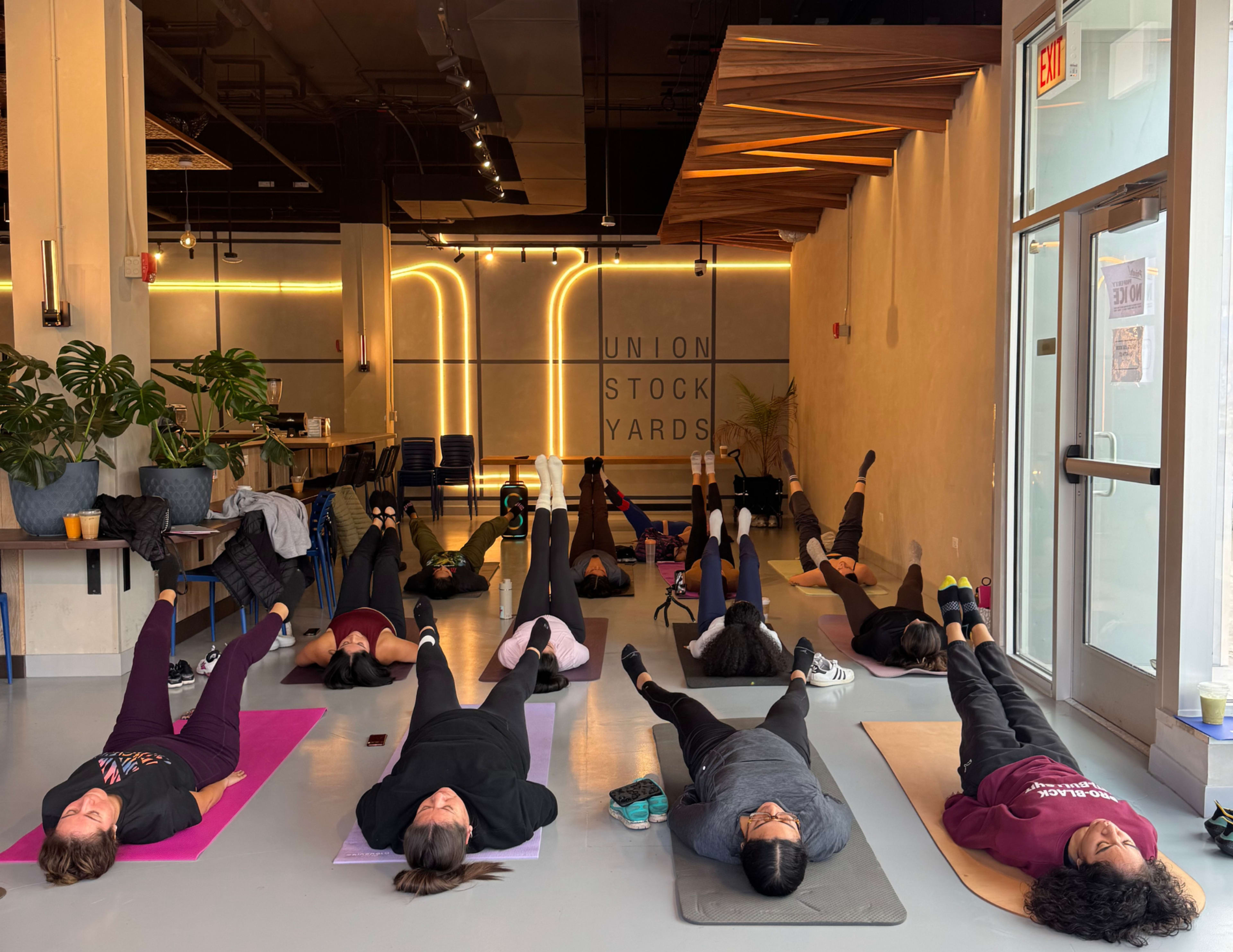 A yoga class is in session with participants lying on mats and performing leg lifts in a brightly lit studio.