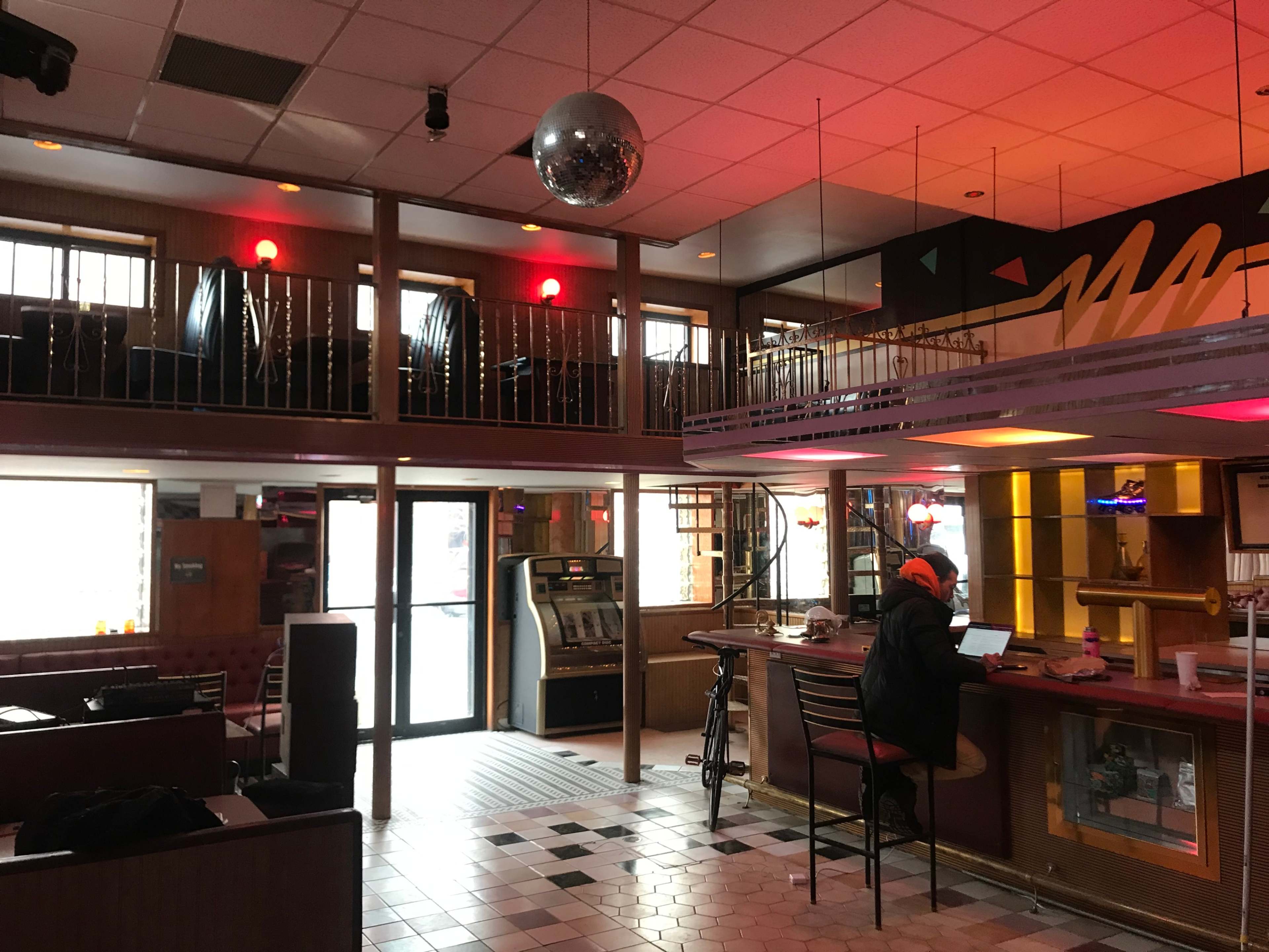 An empty bar area with a person seated at the counter, a disco ball hanging from the ceiling, and booths visible in the background.