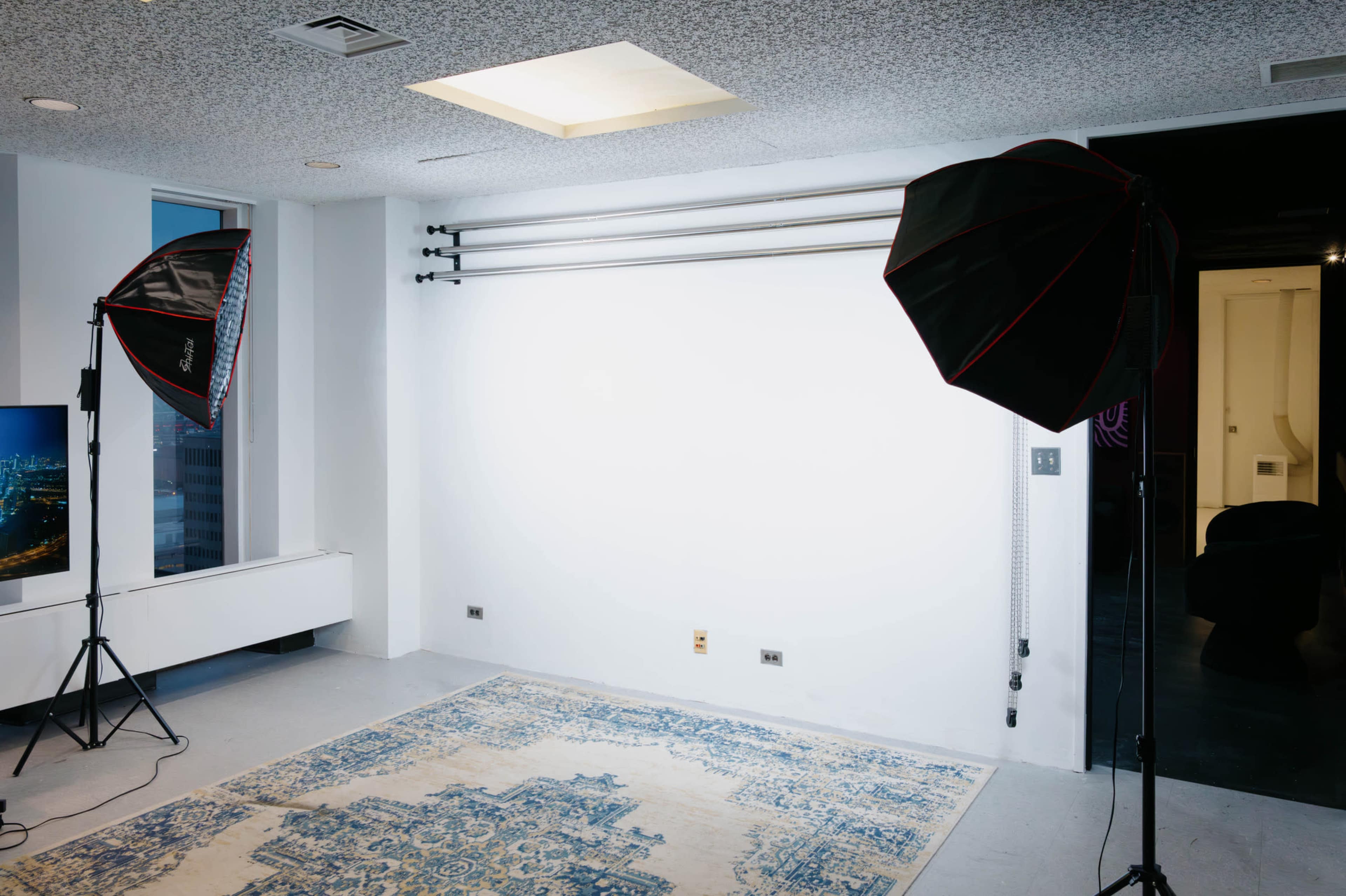 The image shows a well-lit photography studio featuring two large softbox lights positioned on either side of a blank white wall, with a patterned rug on the floor.