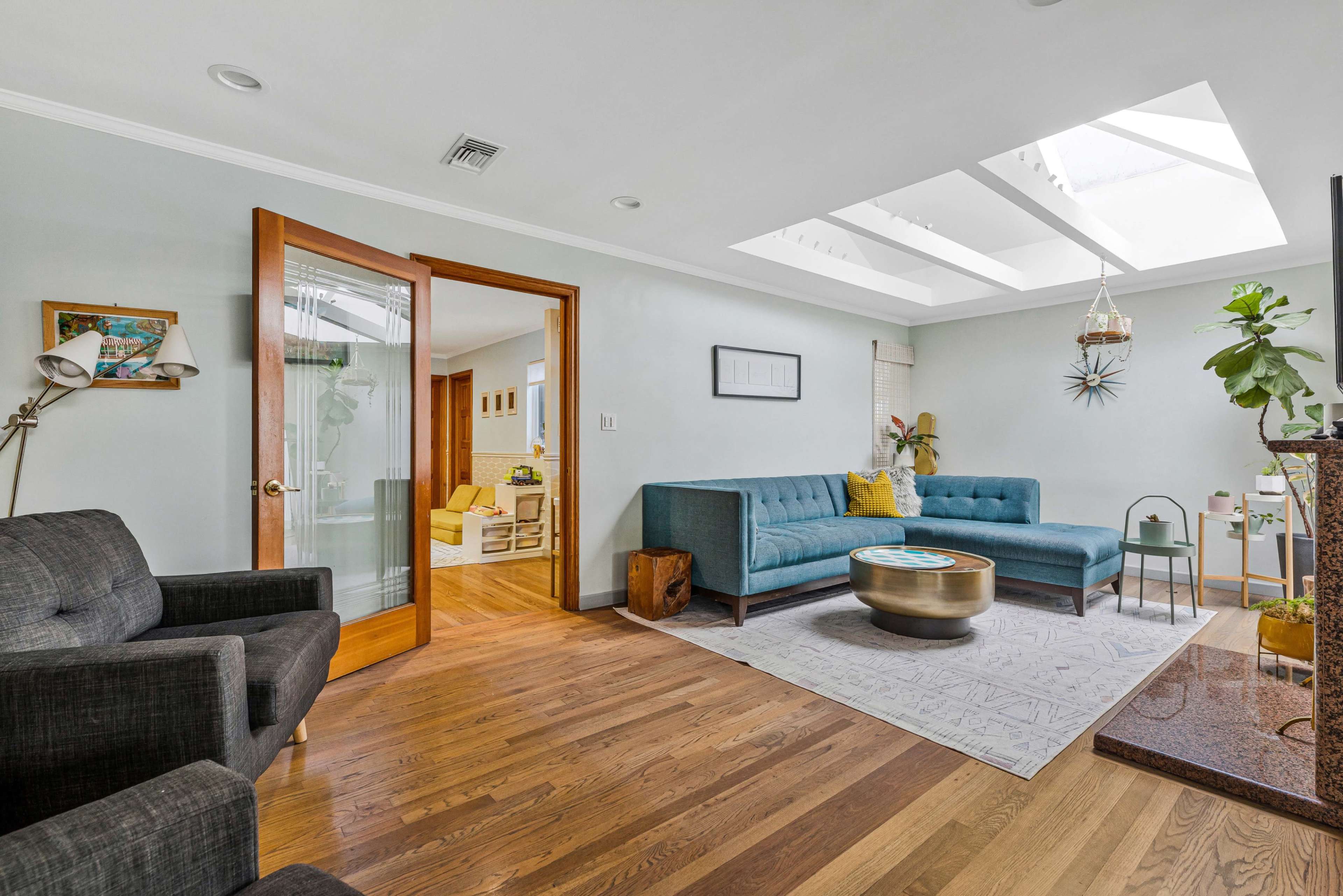 The image shows a living room featuring a teal couch, a wooden coffee table, and large windows that allow natural light to illuminate the space.