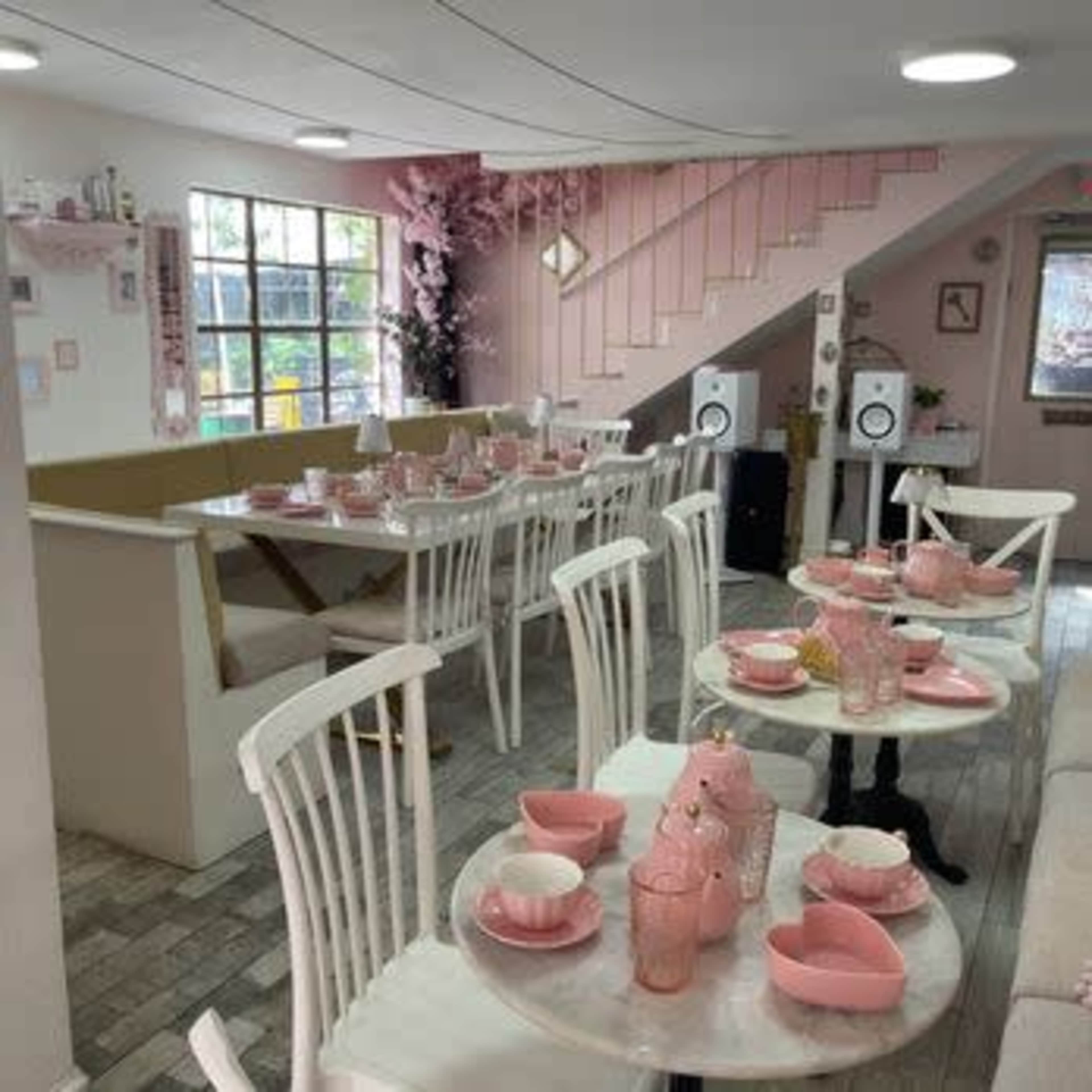The image shows a brightly lit café with white tables and pink tableware, featuring light wood accents and a decorative staircase in the background.