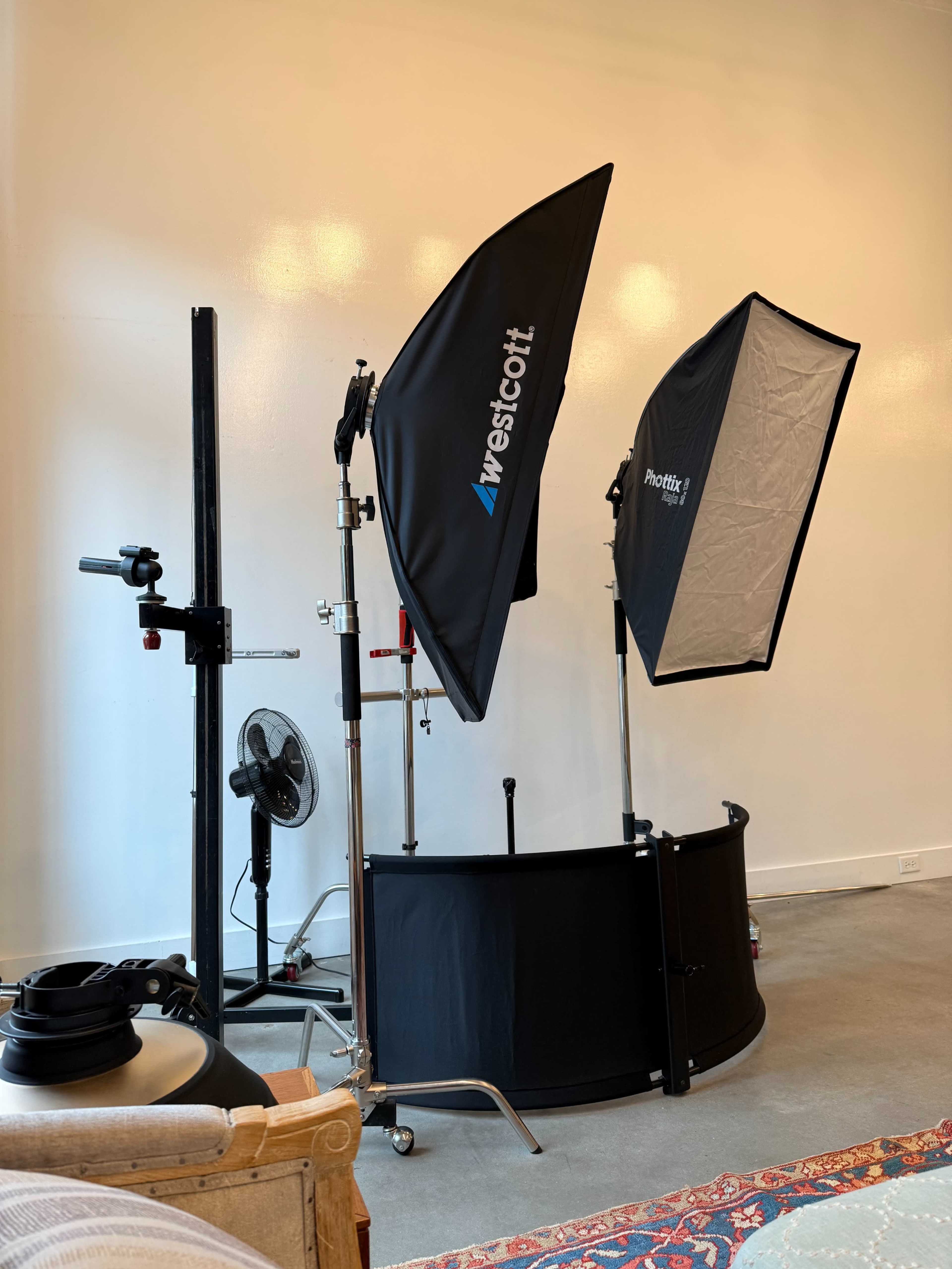 The image shows a photography setup featuring two softbox lights, a backdrop support system, and a fan in a spacious room with a concrete floor.