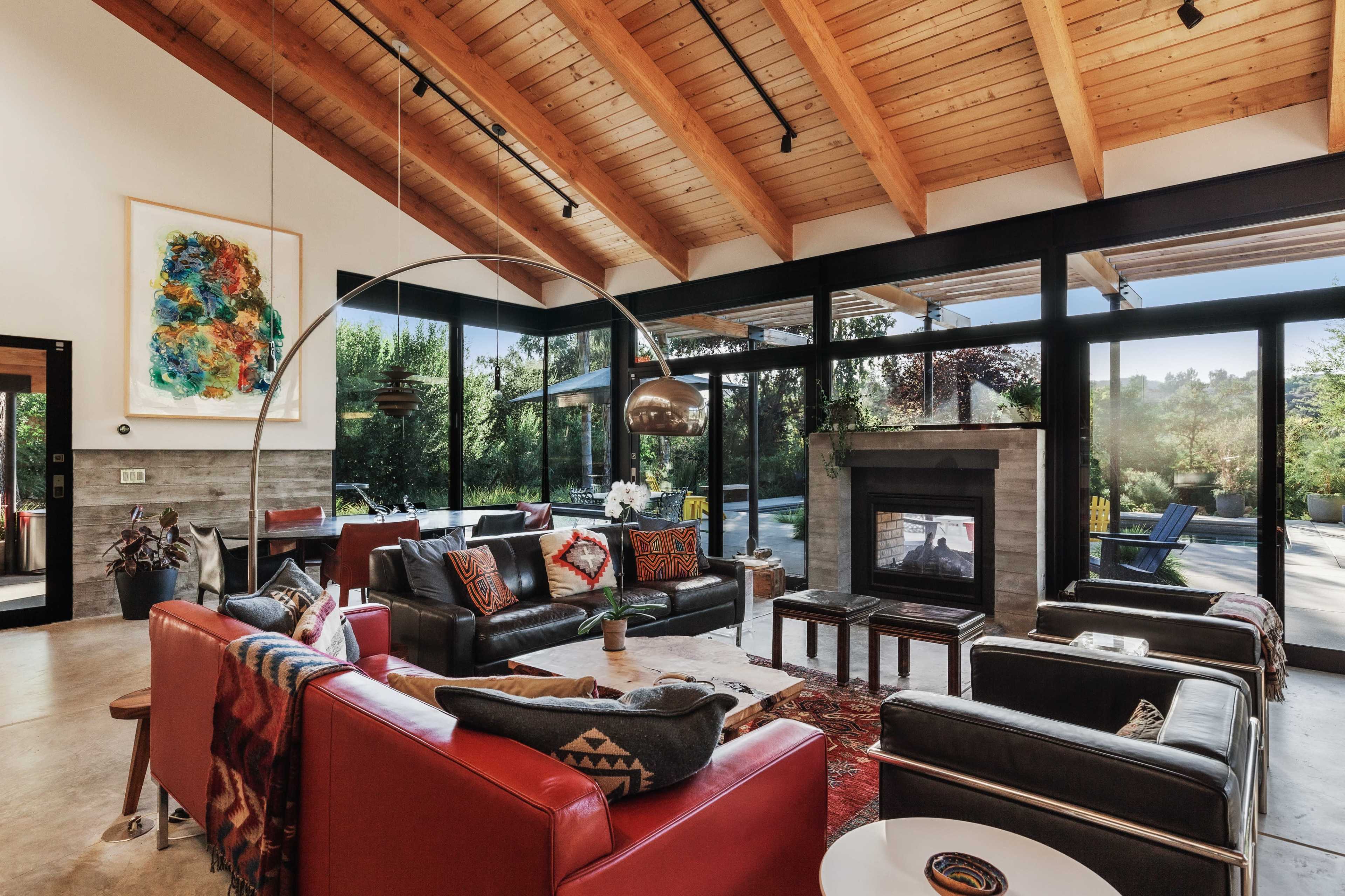 A spacious living room features large glass windows, a modern fireplace, and a mix of comfortable seating arrangements with wooden beams overhead.