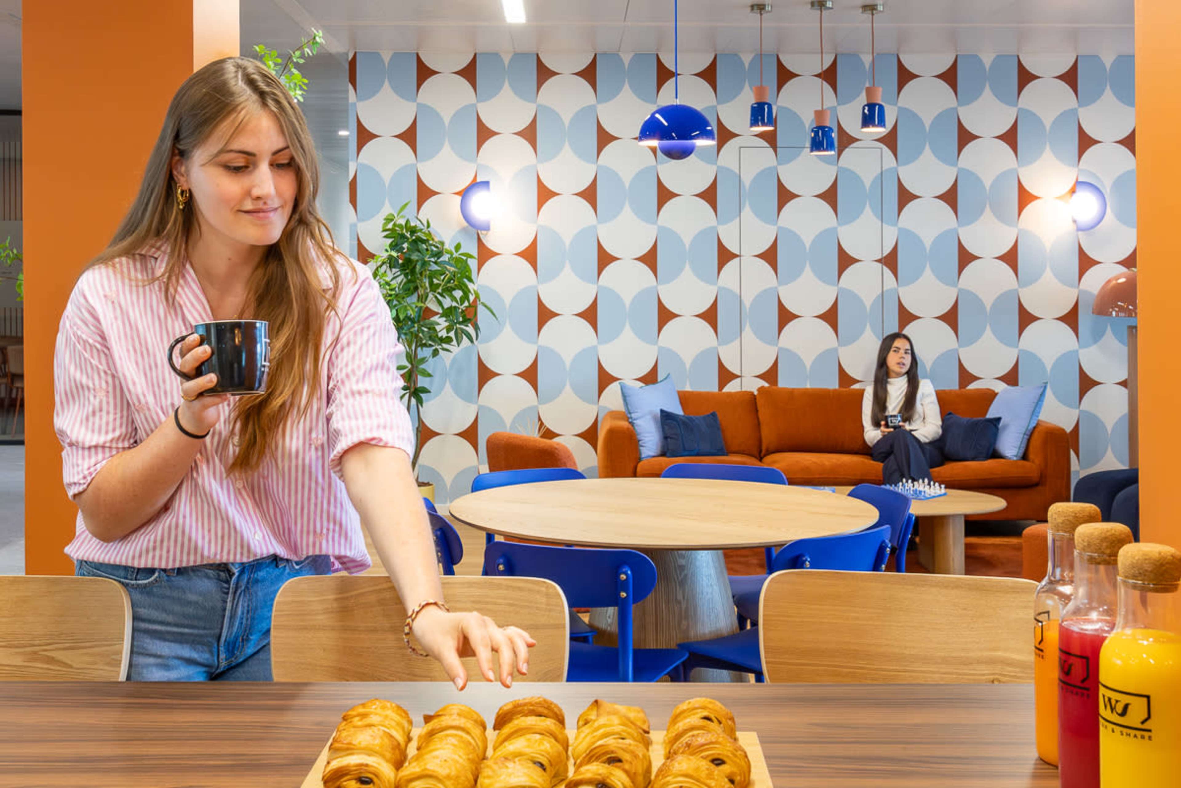 A woman holds a coffee cup while reaching for pastries on a table in a modern café-like space featuring patterned walls and colorful seating.