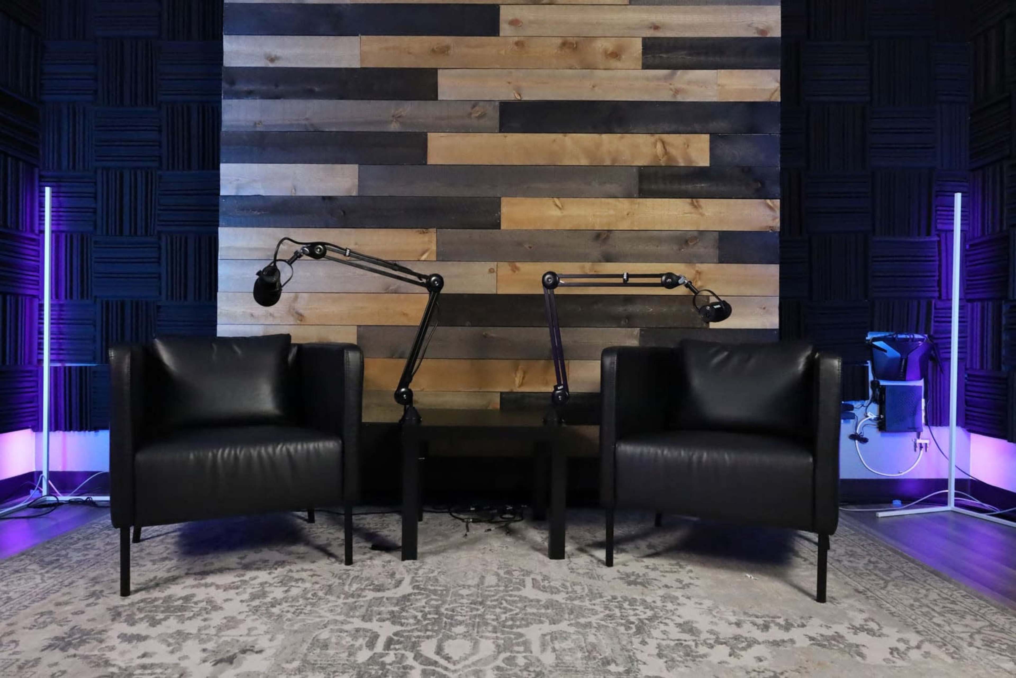 Two black chairs positioned in front of a wooden wall, with microphone arms extended on either side and purple ambient lighting.