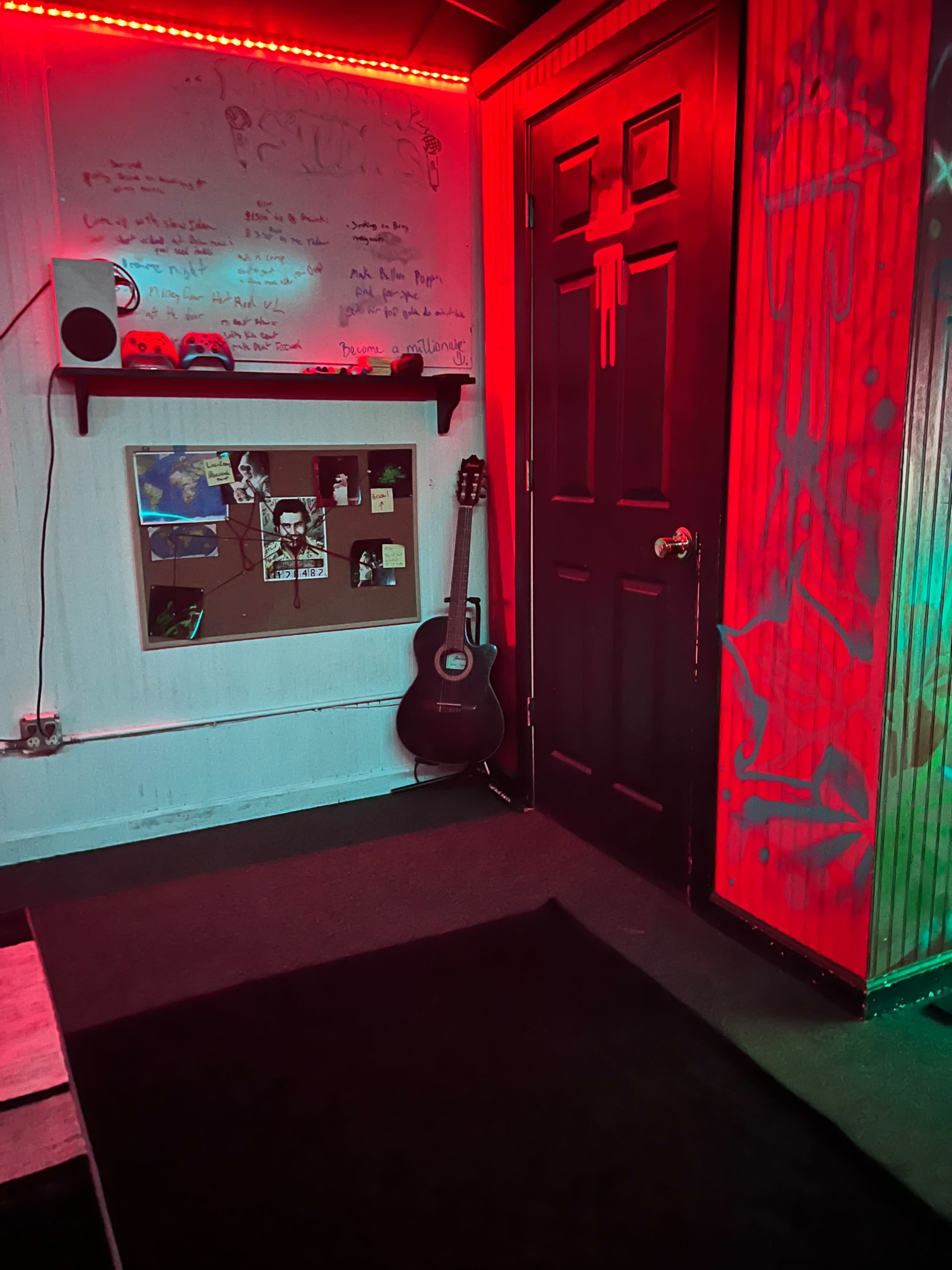 The image shows a dimly lit room with red neon lighting, featuring a decorated wall, a black door, a guitar leaning against the wall, and a shelf with various items above a makeshift carpet.
