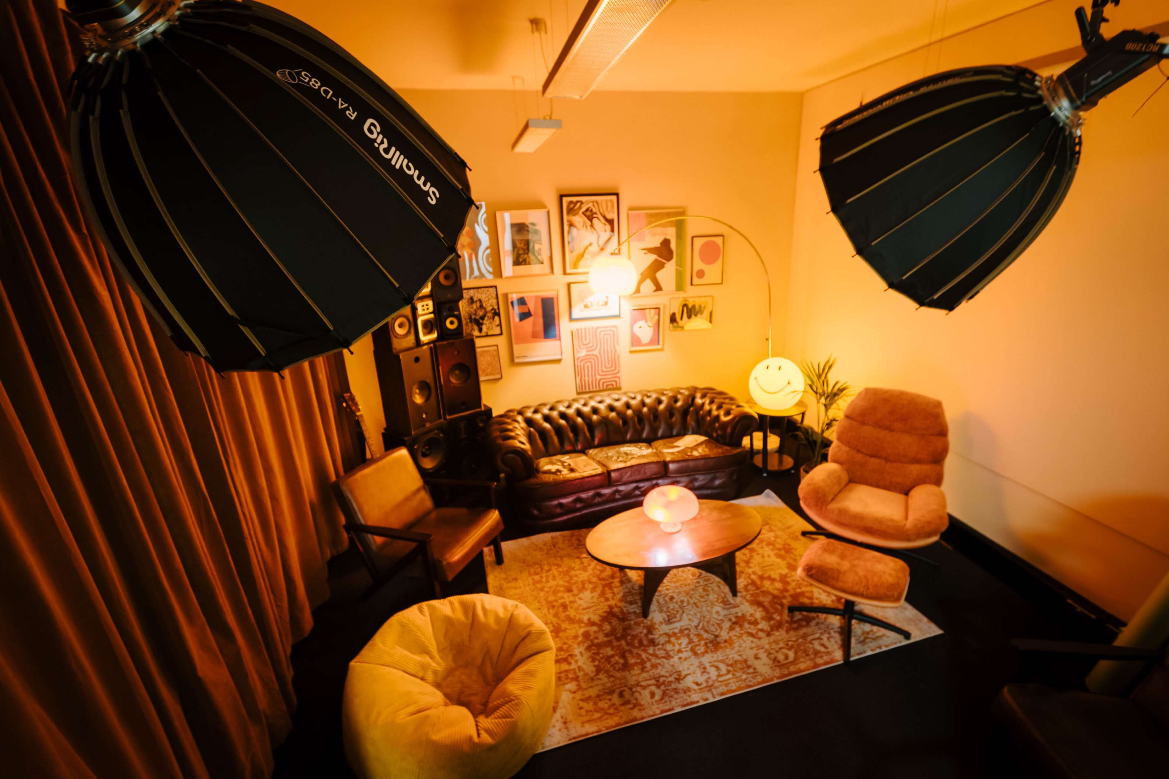 The image shows a cozy lounge area with a leather sofa, a circular table, and various decorative elements, illuminated by large softbox lights.