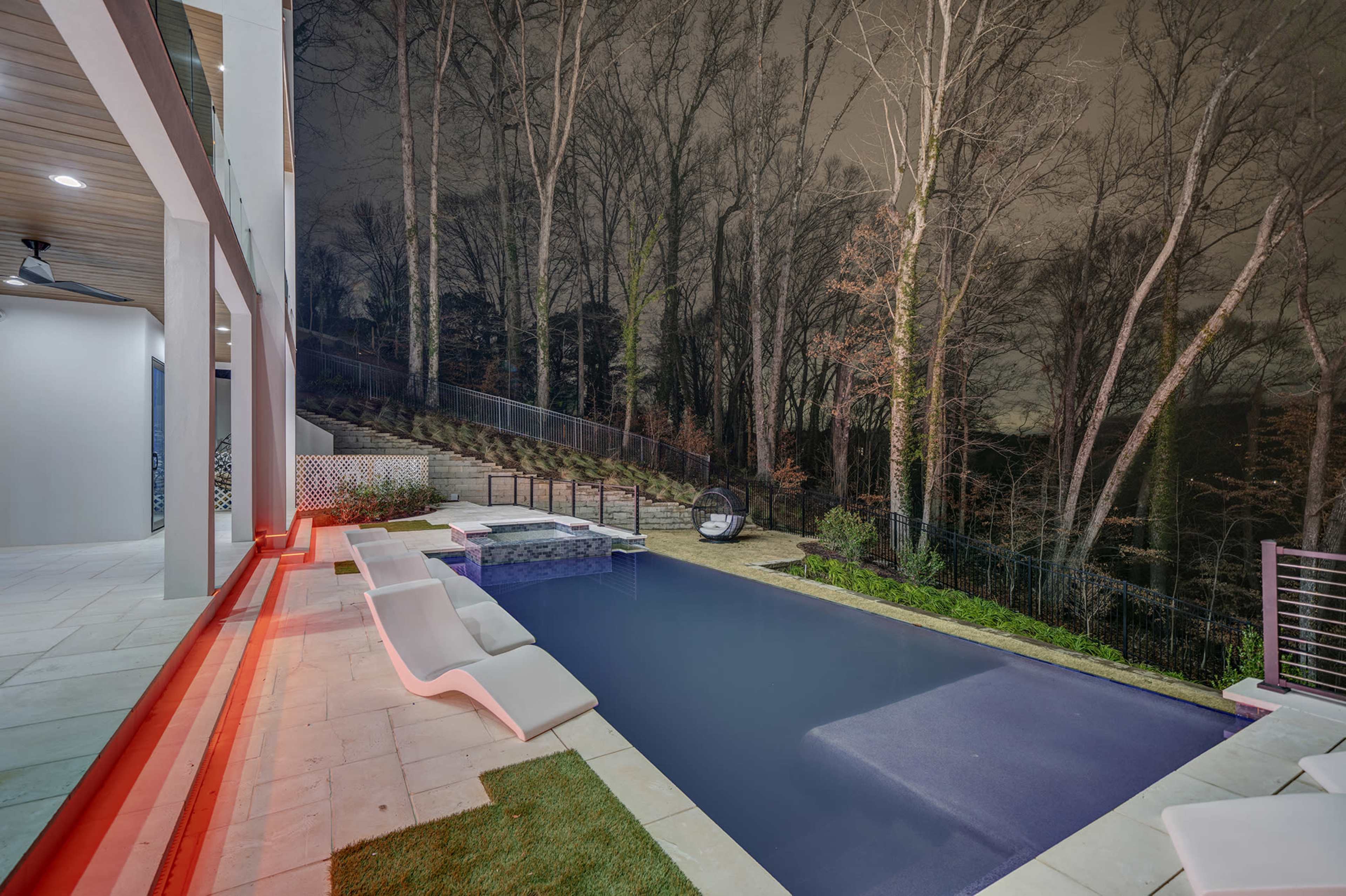 A rectangular swimming pool is situated next to a modern deck area, surrounded by trees in a dimly lit setting.
