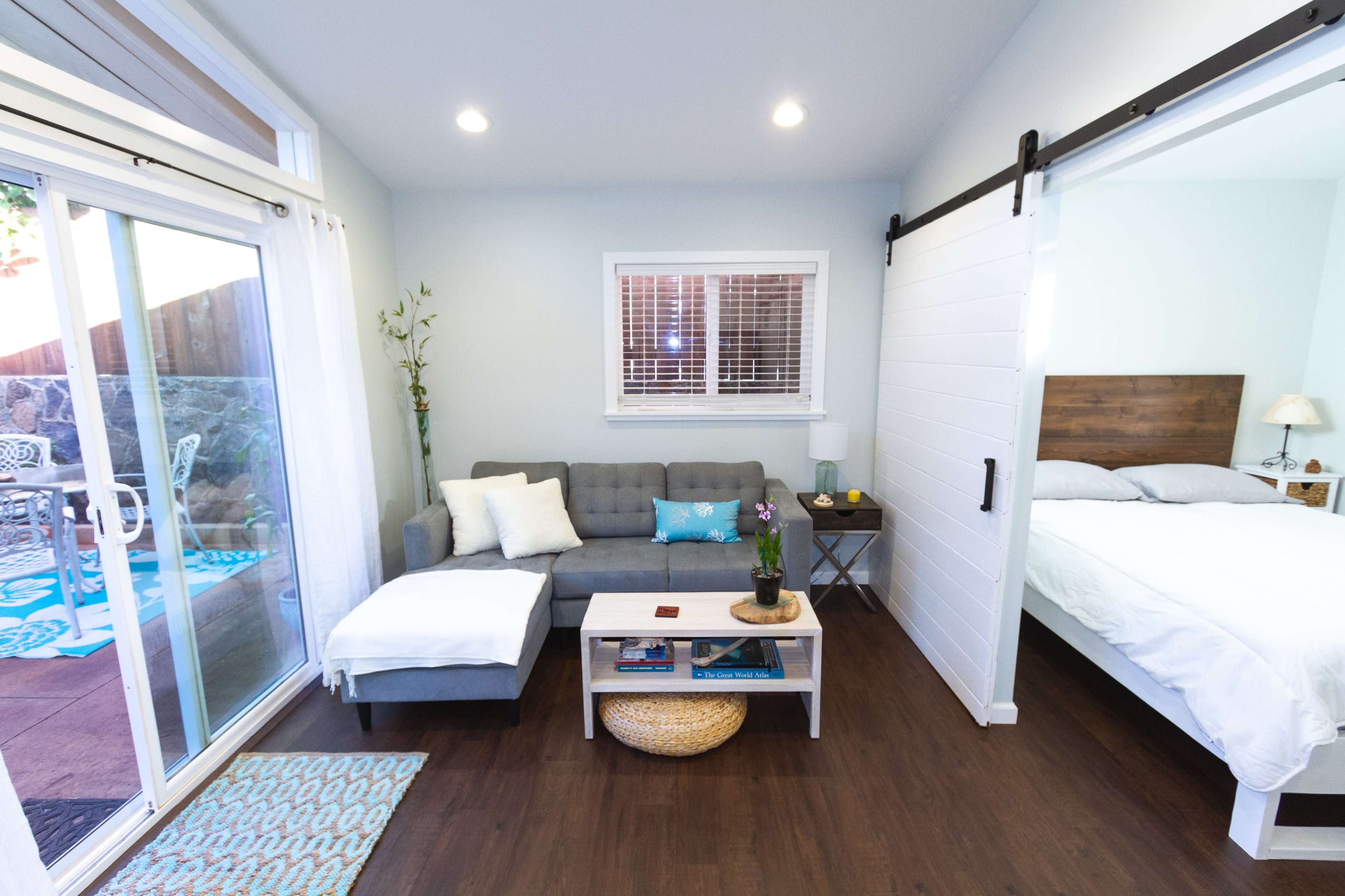 A small, well-lit living area with a gray sofa, a coffee table, and a bedroom area separated by a sliding door.