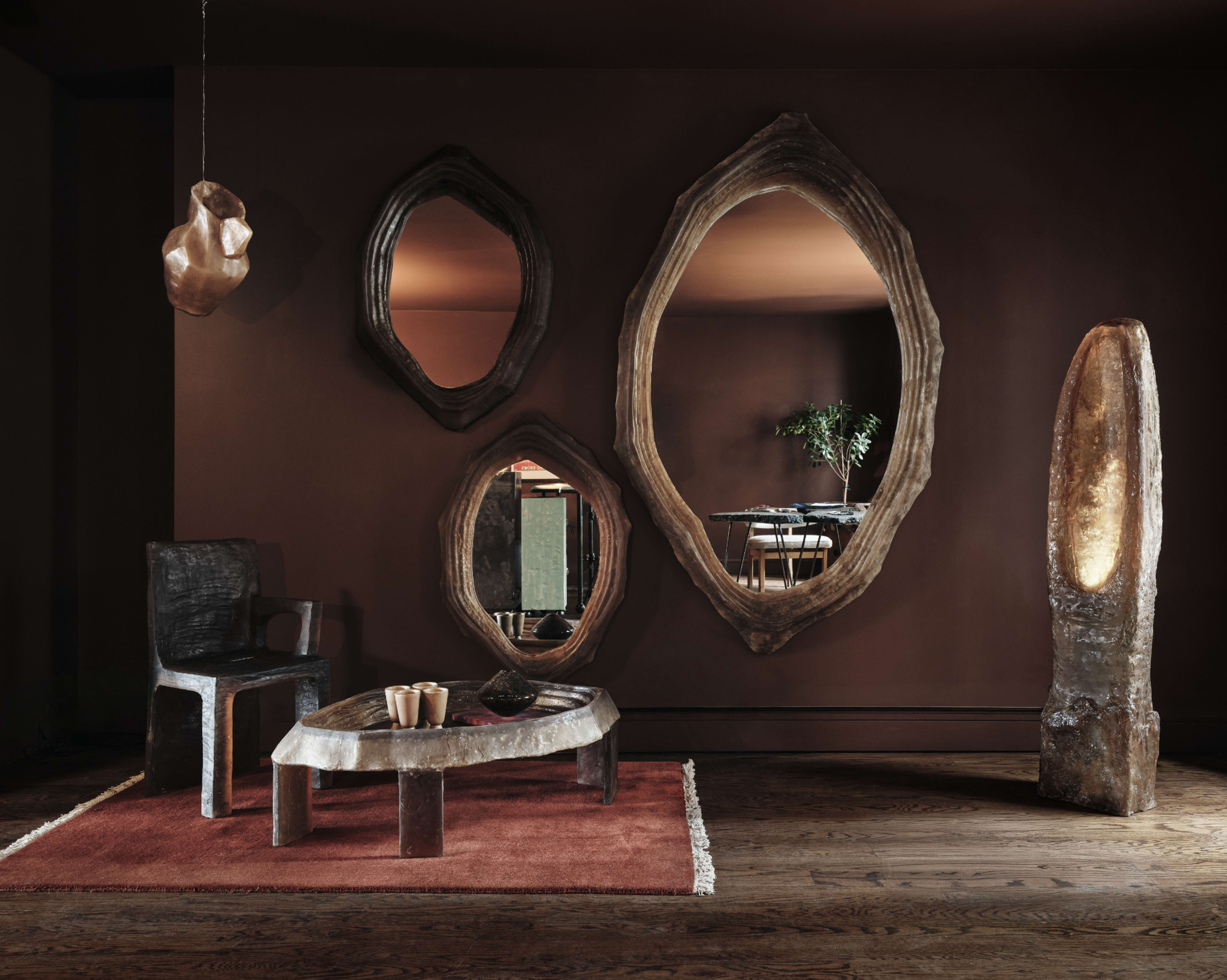 The image shows a modern interior with several organic-shaped wooden mirrors mounted on a dark wall, a rustic table and chair, and a decorative stone lamp, all set on a warm-toned area rug.