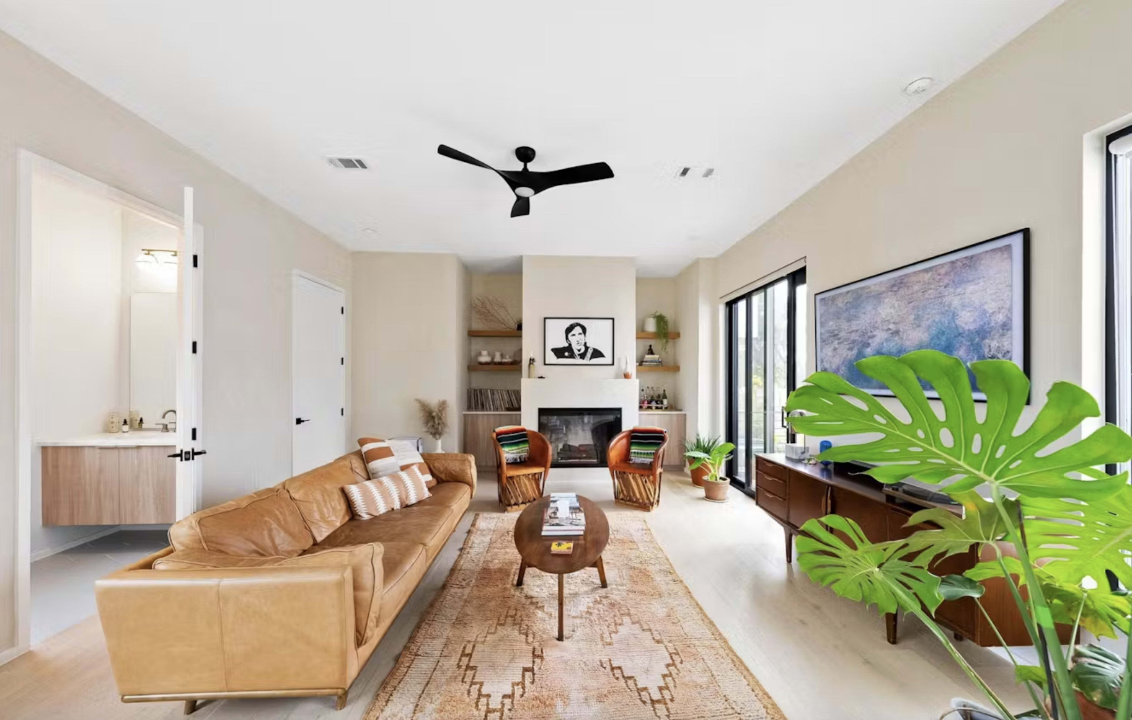 The living room features a tan leather sofa, a large rug with intricate patterns, a wall-mounted television, and a ceiling fan, with natural light streaming through large windows.