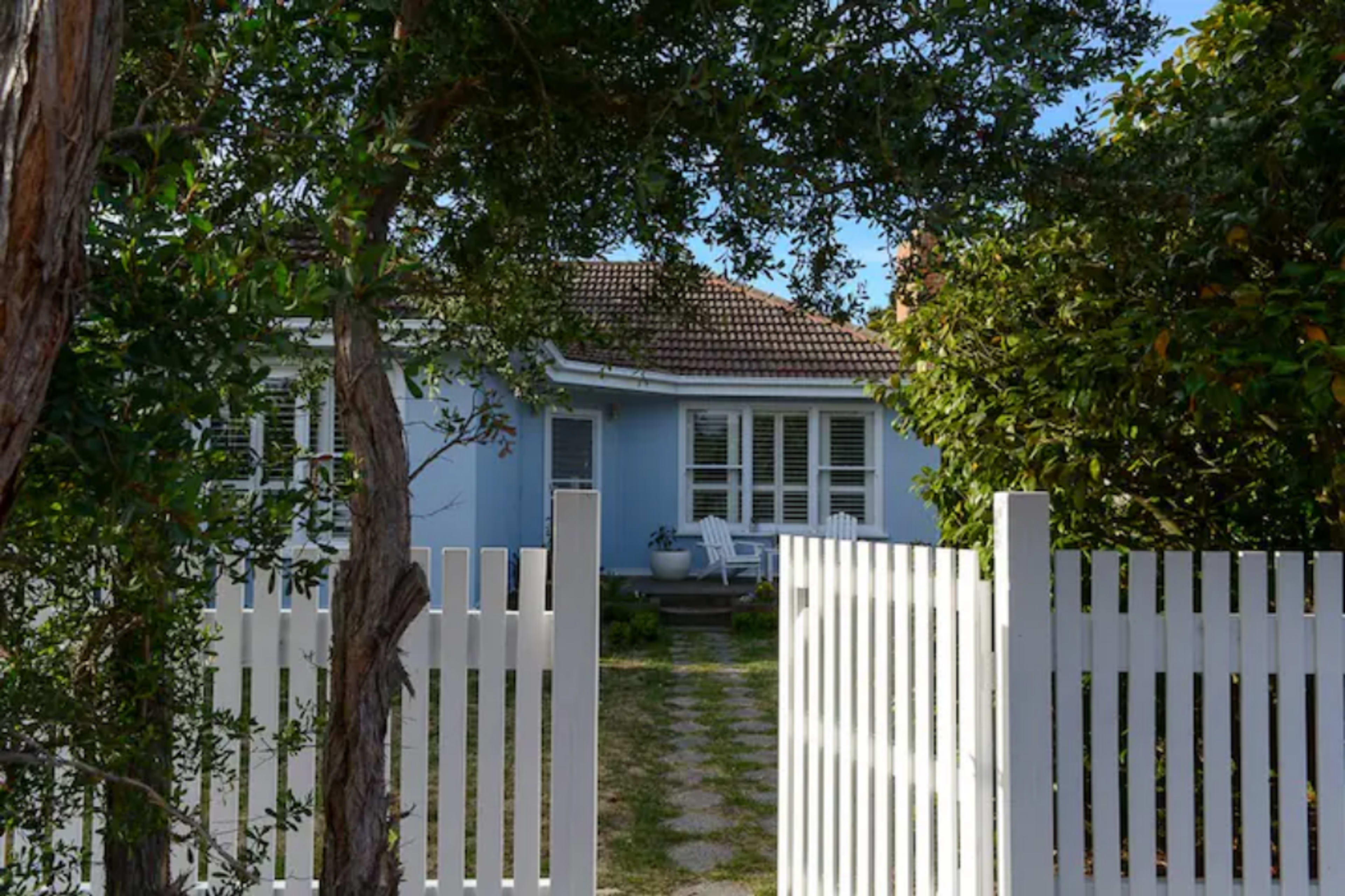A white picket fence frames a pathway leading to a light blue house with a tiled roof and a front porch.