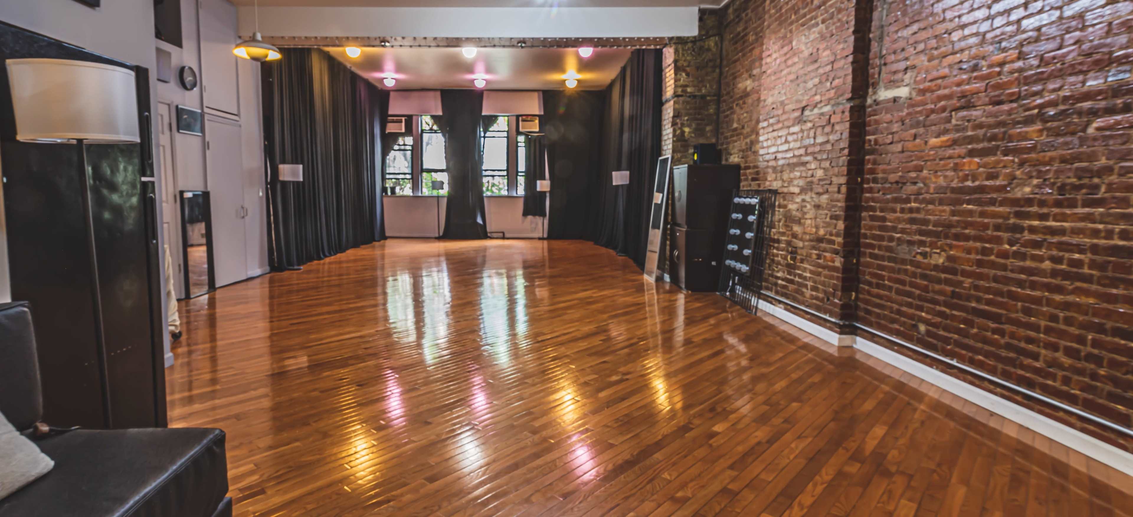 A spacious room with polished wooden floors, exposed brick walls, and curtains at the far end, illuminated by overhead lights.