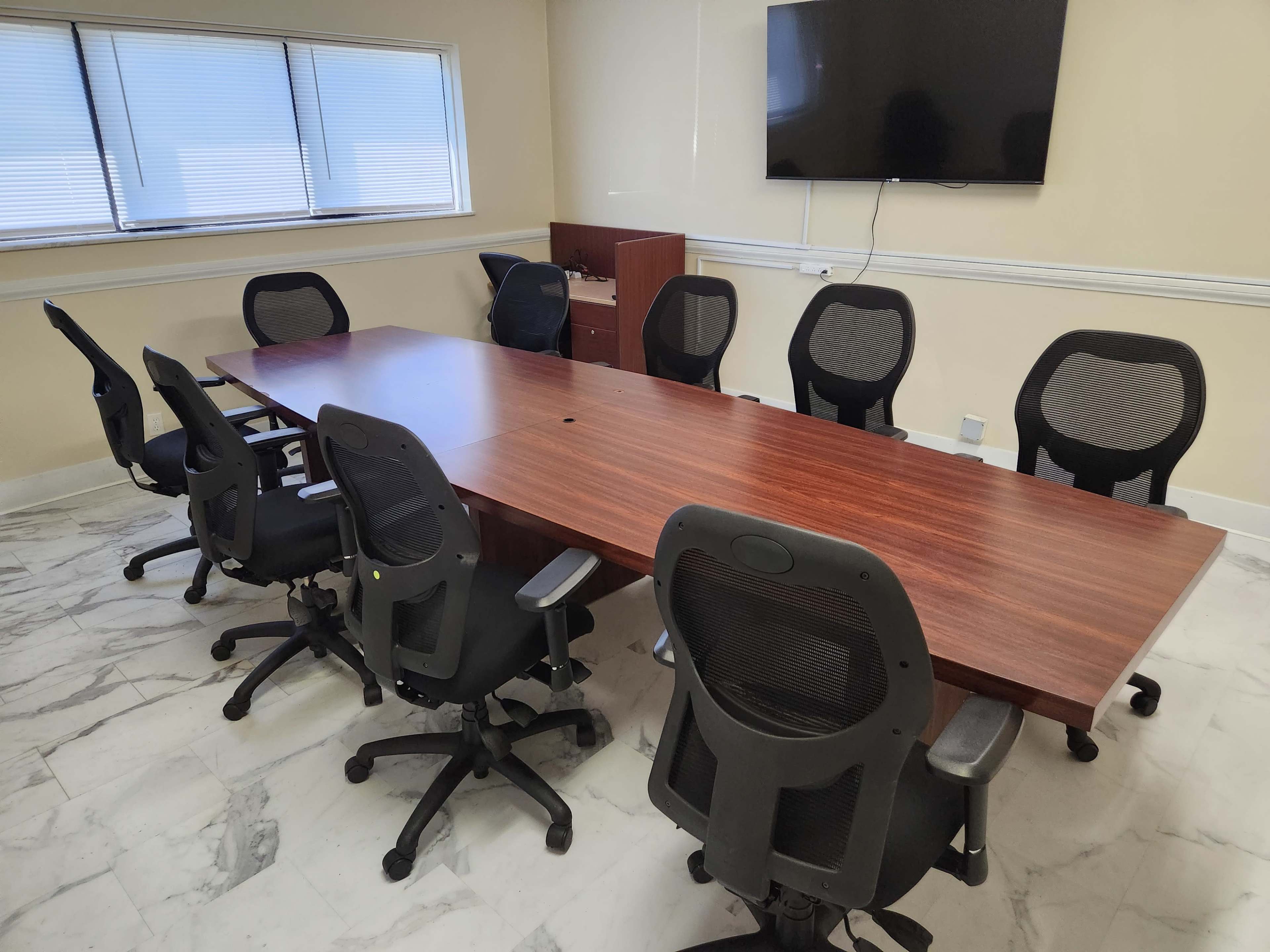 A large conference table with eight ergonomic chairs is positioned in a well-lit meeting room featuring a wall-mounted television.