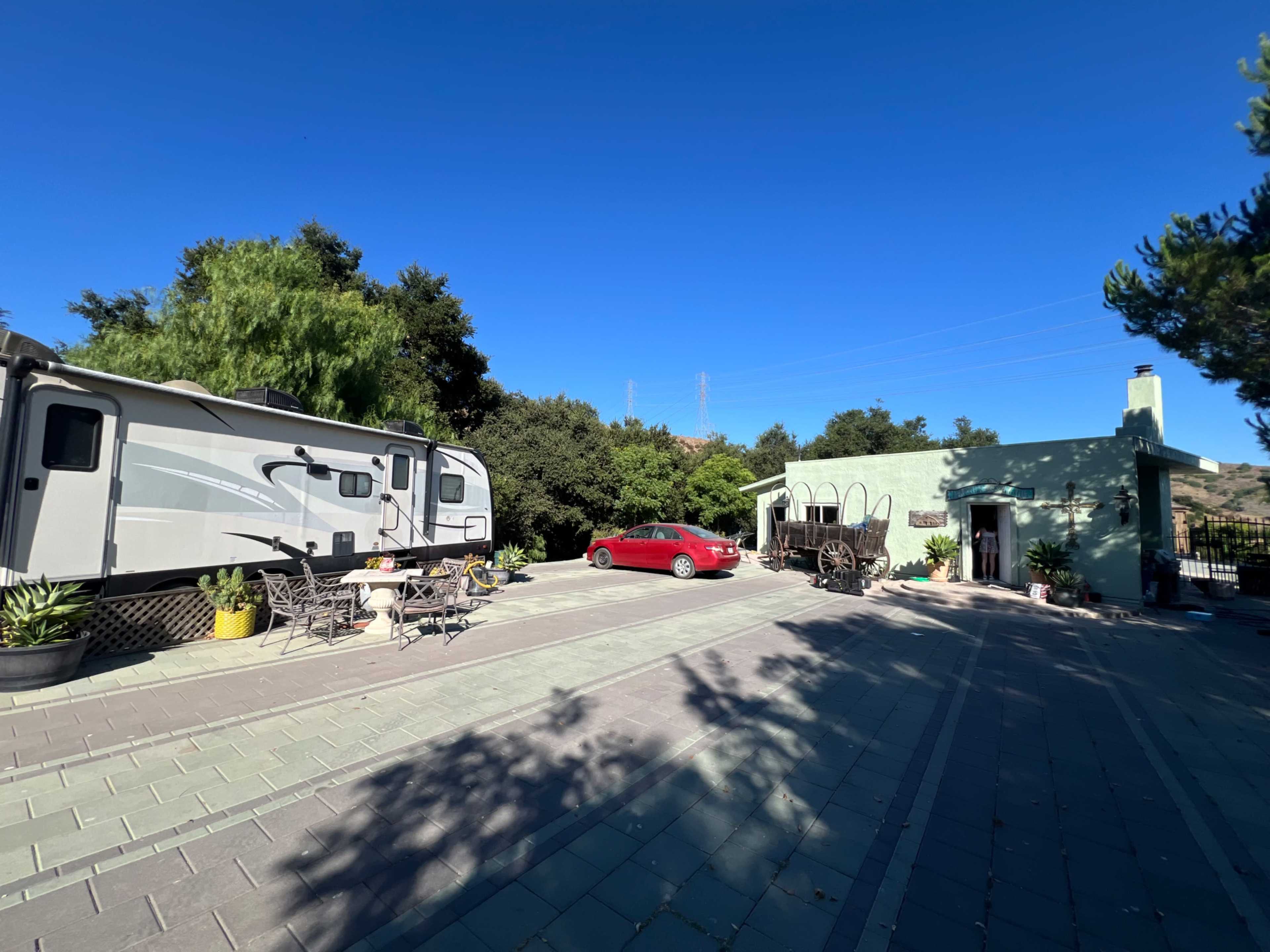 A recreational vehicle is parked beside a red car in front of a small green building, surrounded by trees and under a clear blue sky.