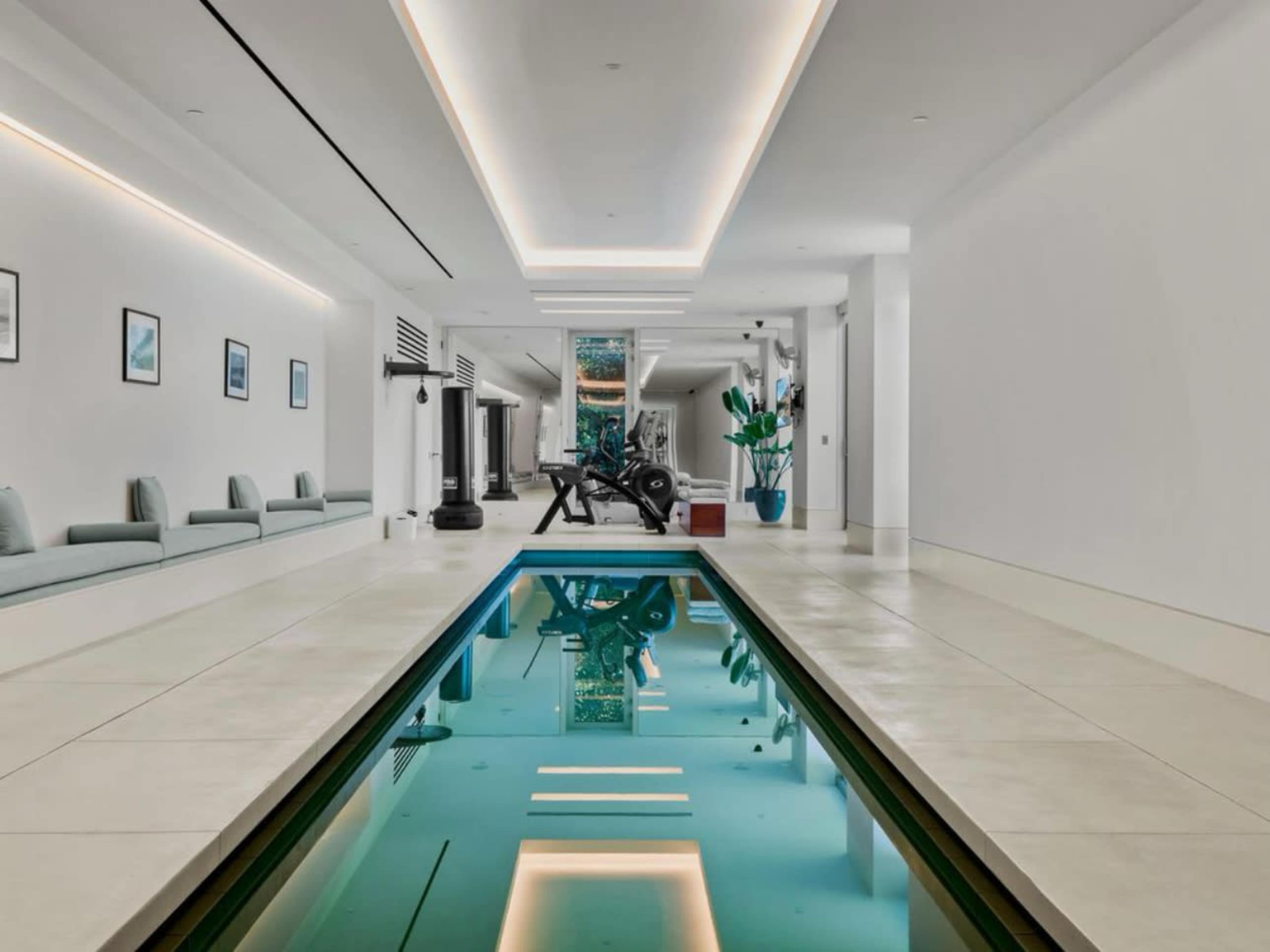 A modern indoor swimming pool surrounded by white walls, minimalist furniture, and exercise equipment in a spacious room.