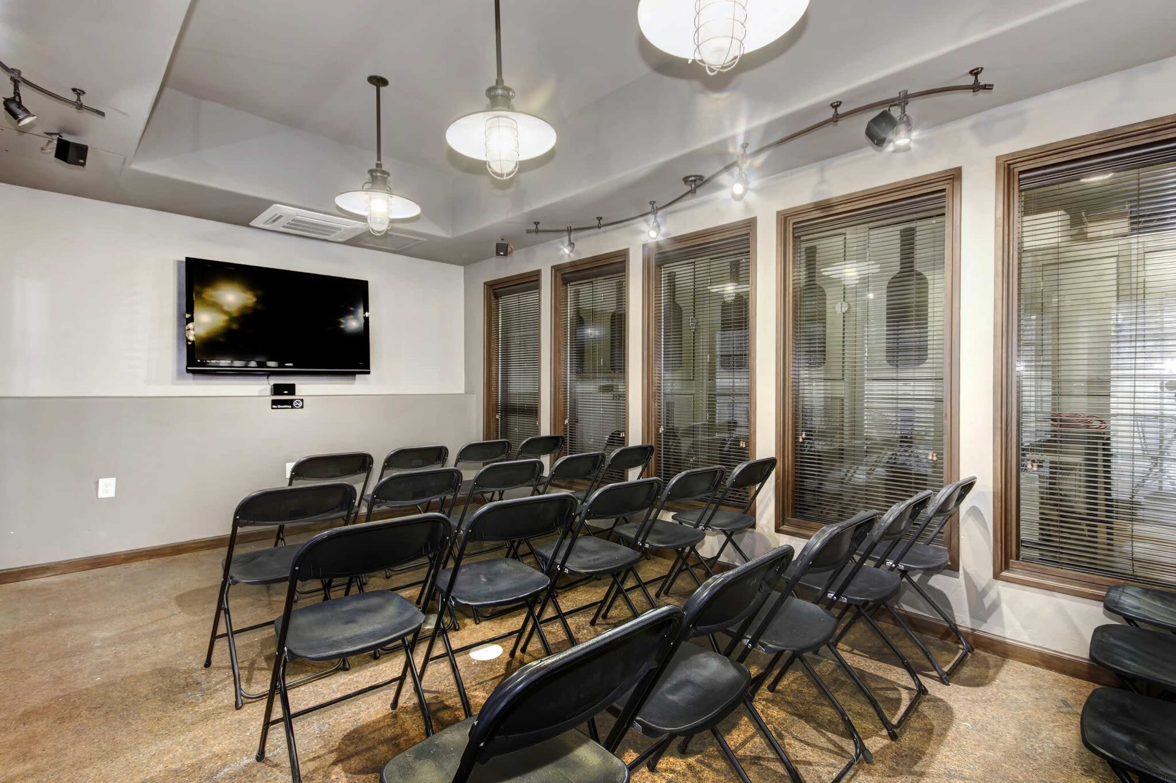 A small meeting room with several rows of black folding chairs facing a wall-mounted television and large windows with blinds.