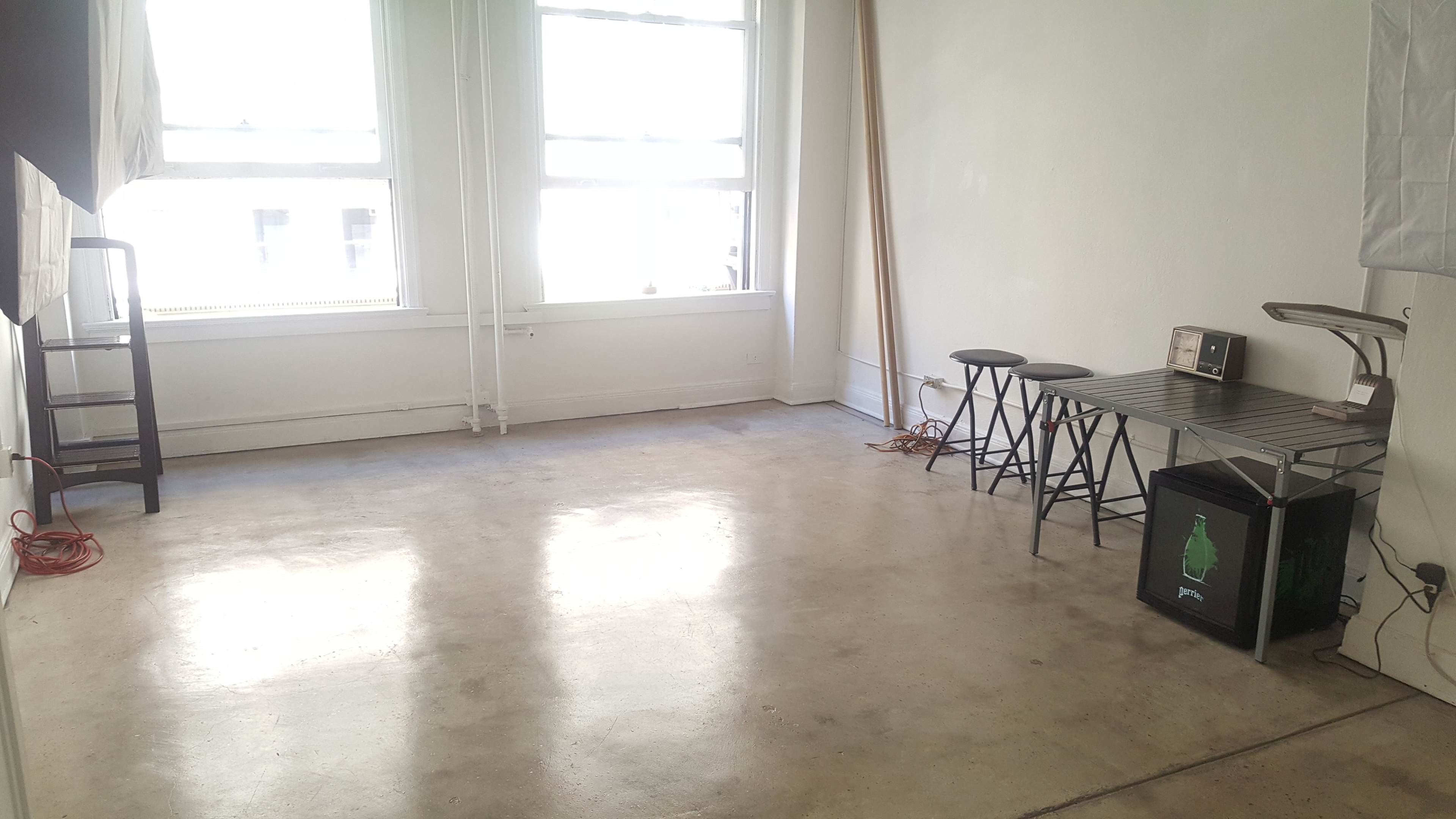 A minimalist room with a concrete floor, two large windows, a small kitchen table, three stools, a microwave, and a compact fridge.