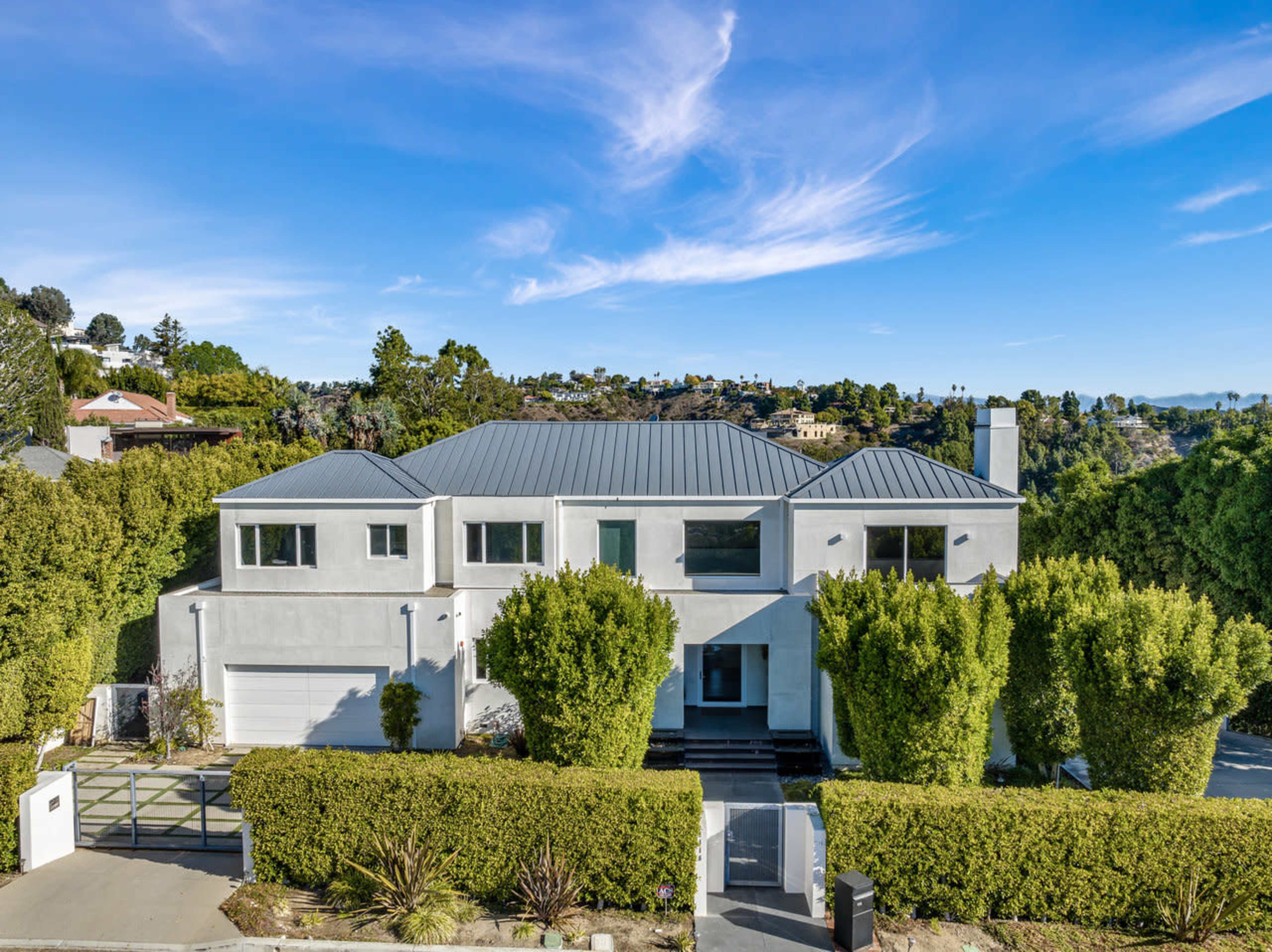 A two-story gray house with a metal roof is surrounded by neatly trimmed hedges and trees on a hillside.