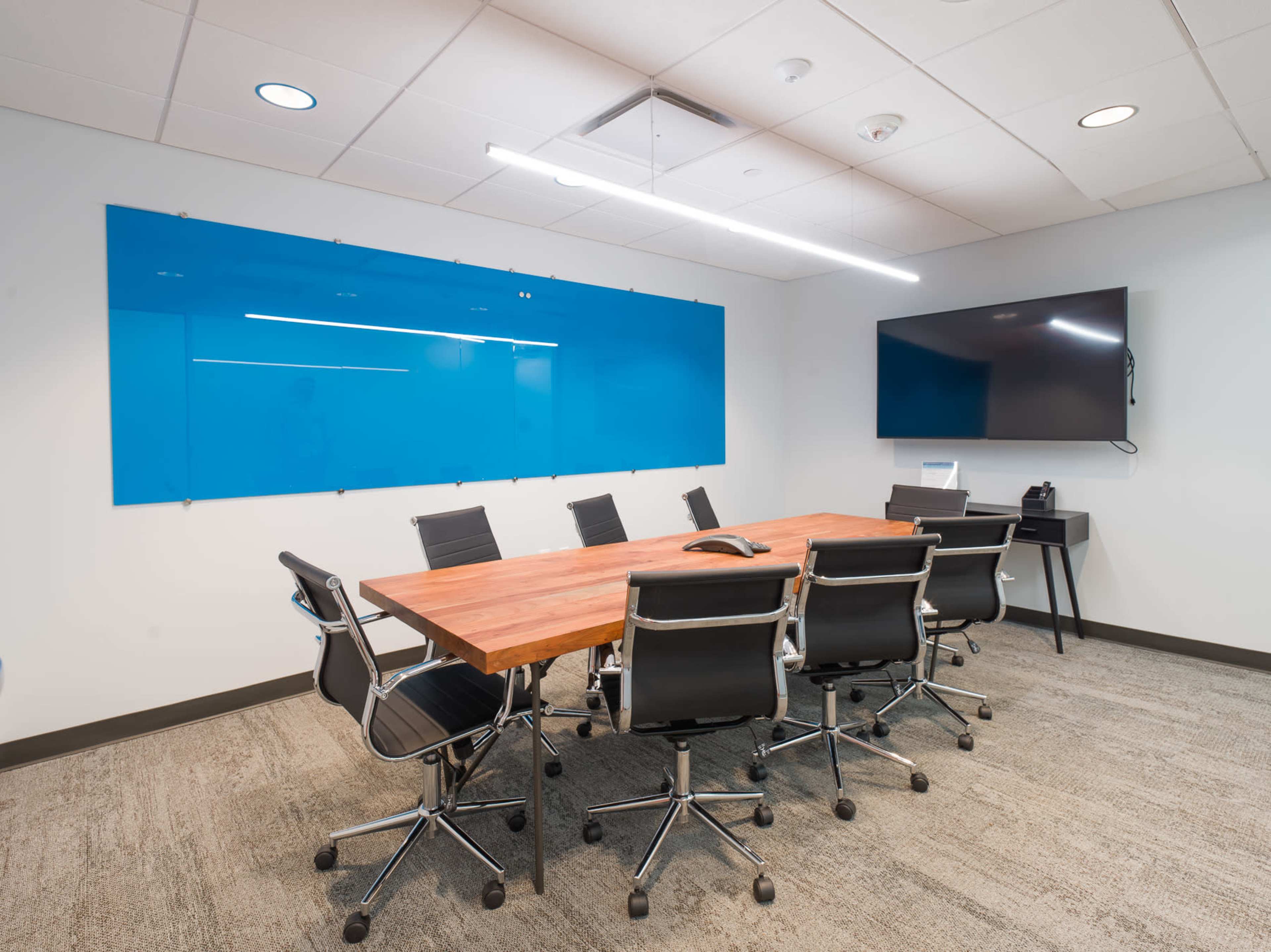 A modern conference room features a large wooden table surrounded by black swivel chairs, with a blue glass wall and a wall-mounted television.