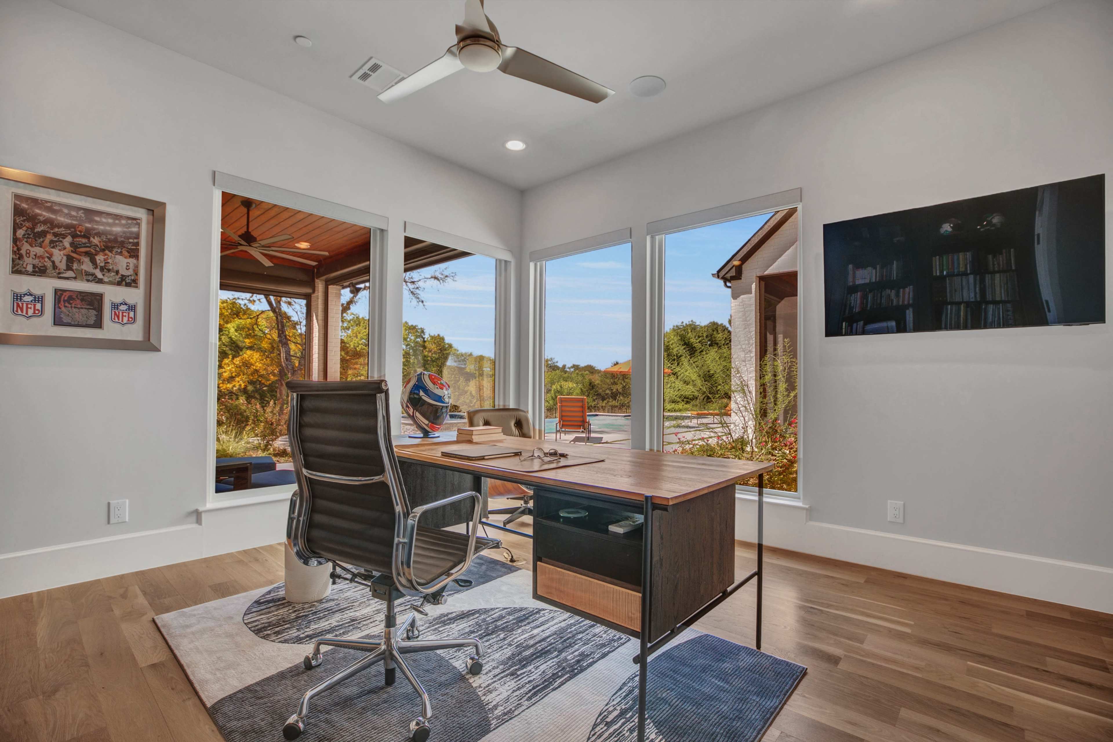 A modern home office features a wooden desk, ergonomic chair, and large windows overlooking a landscaped outdoor area.