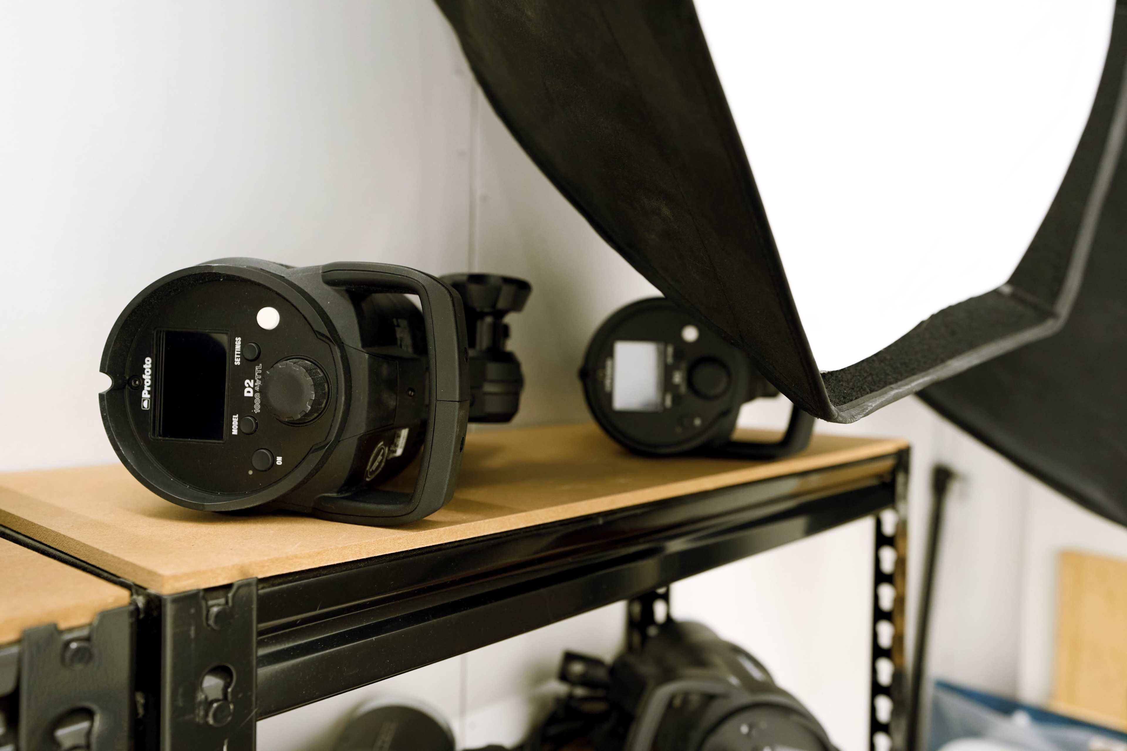 The image shows studio equipment, including a camera flash unit and a softbox, arranged on a shelf in a photography workspace.