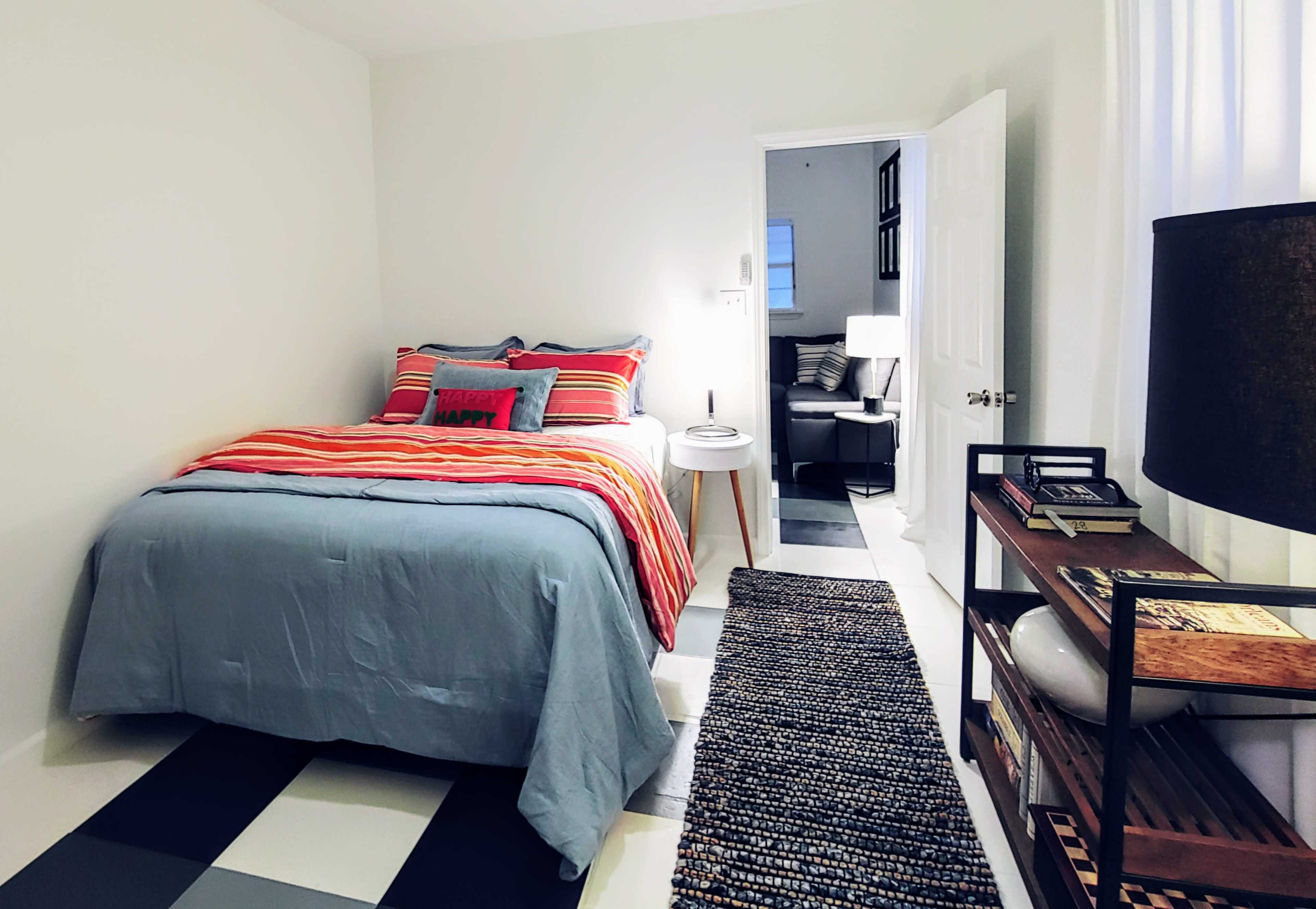 A neatly arranged bedroom features a bed with striped bedding, a side table, a lamp, and a bookshelf alongside a doorway.