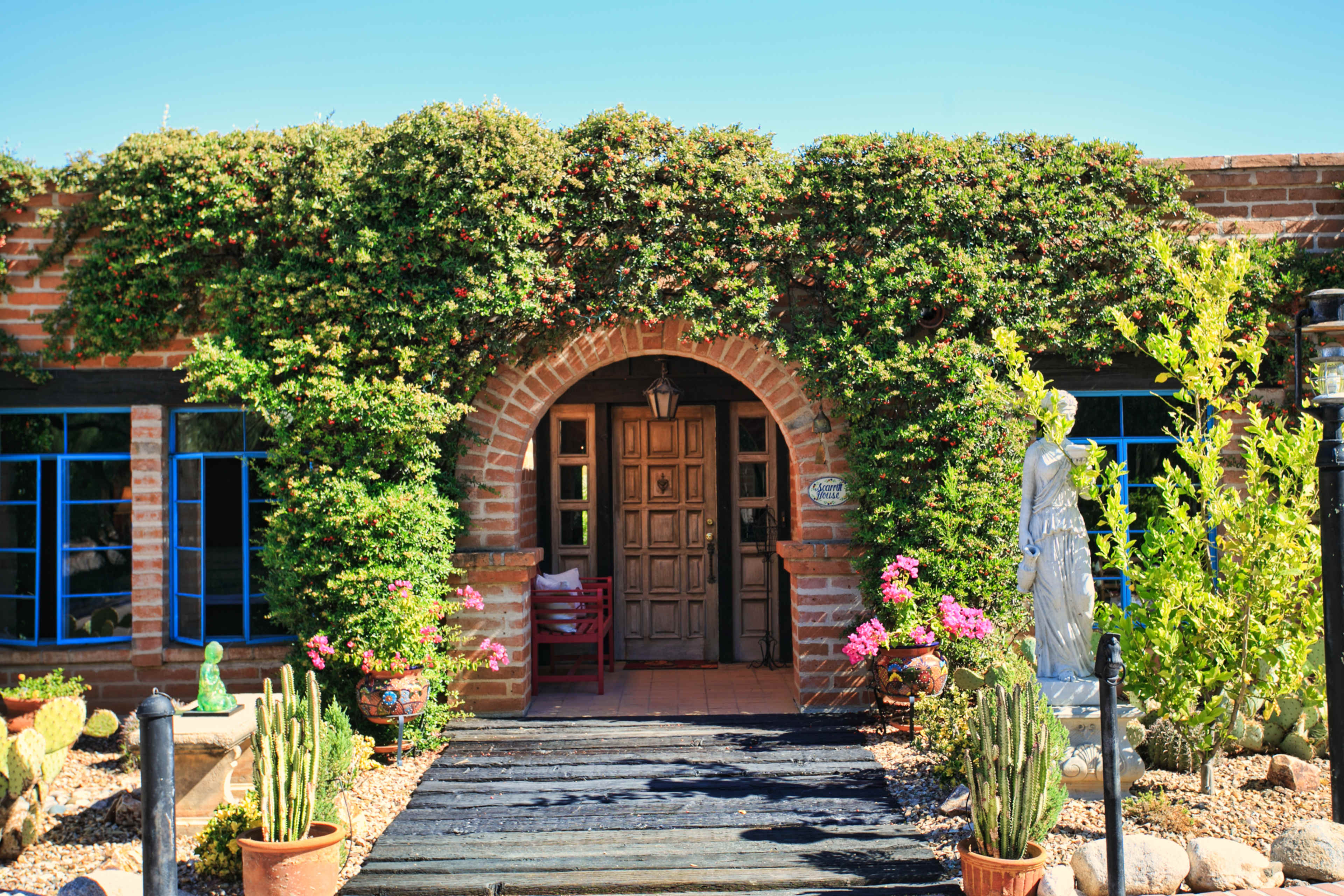 A brick building with a wooden door, surrounded by lush greenery and potted plants, featuring a stone statue and blue window frames.