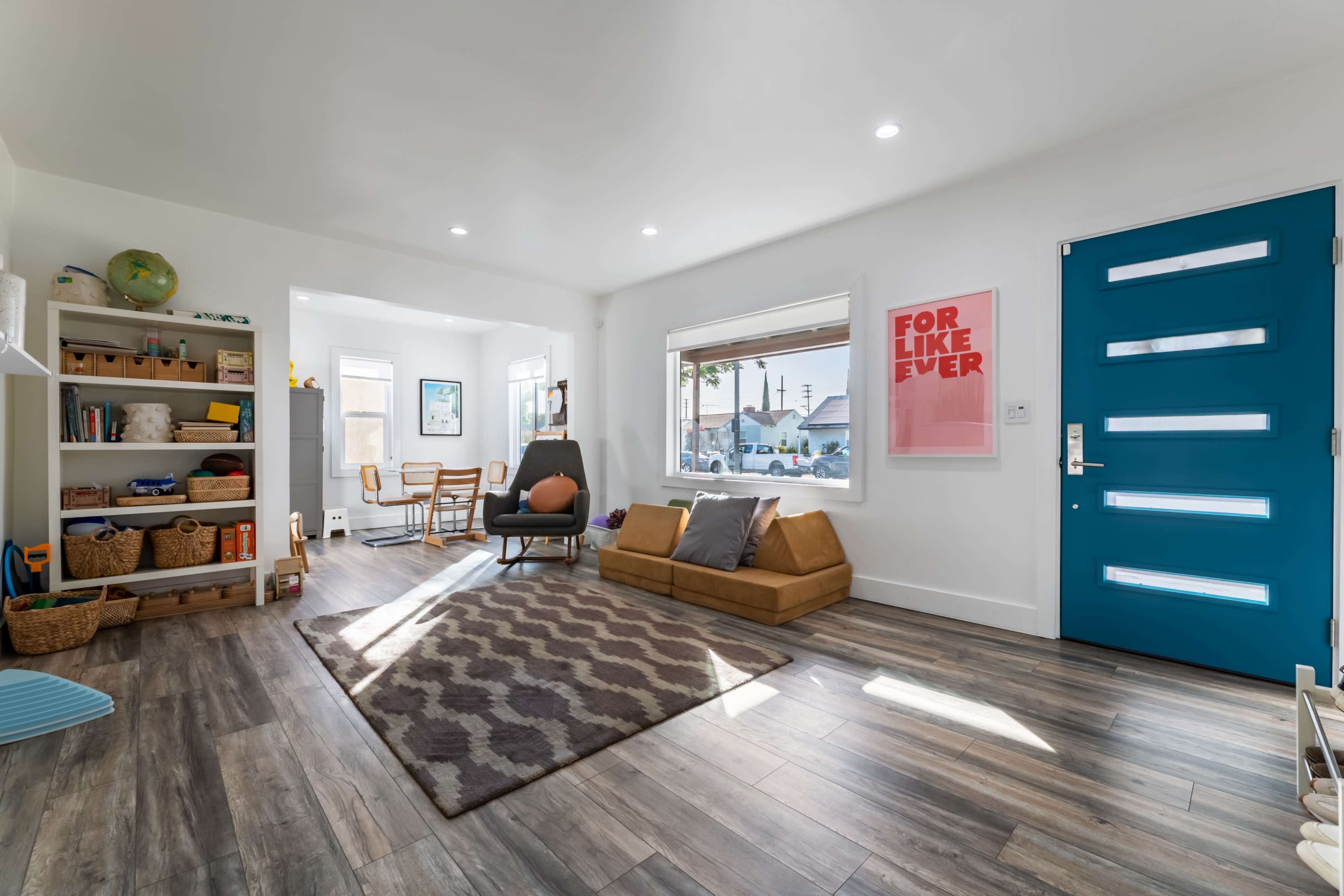 A bright living room features a blue front door, a brown sofa, a patterned rug, and a small dining area with wooden chairs.