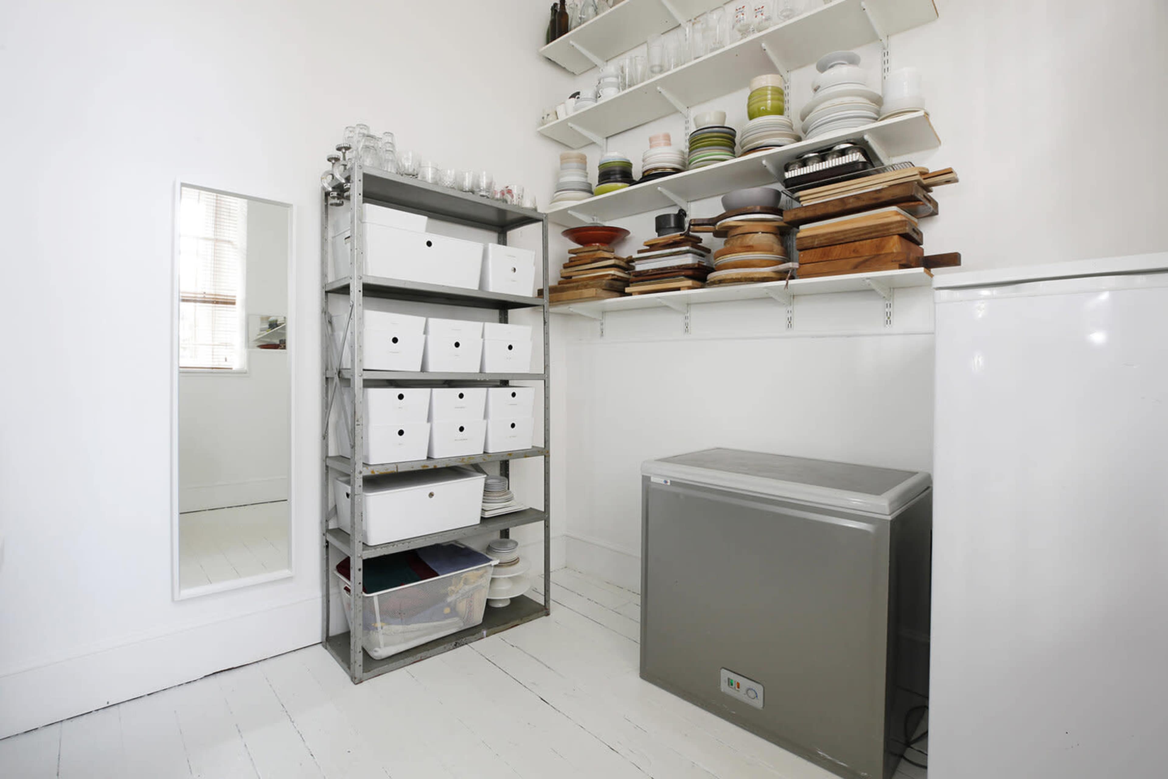 A clean storage area with shelves holding dishes and wooden boards, alongside a metal shelving unit filled with boxes and a chest freezer.