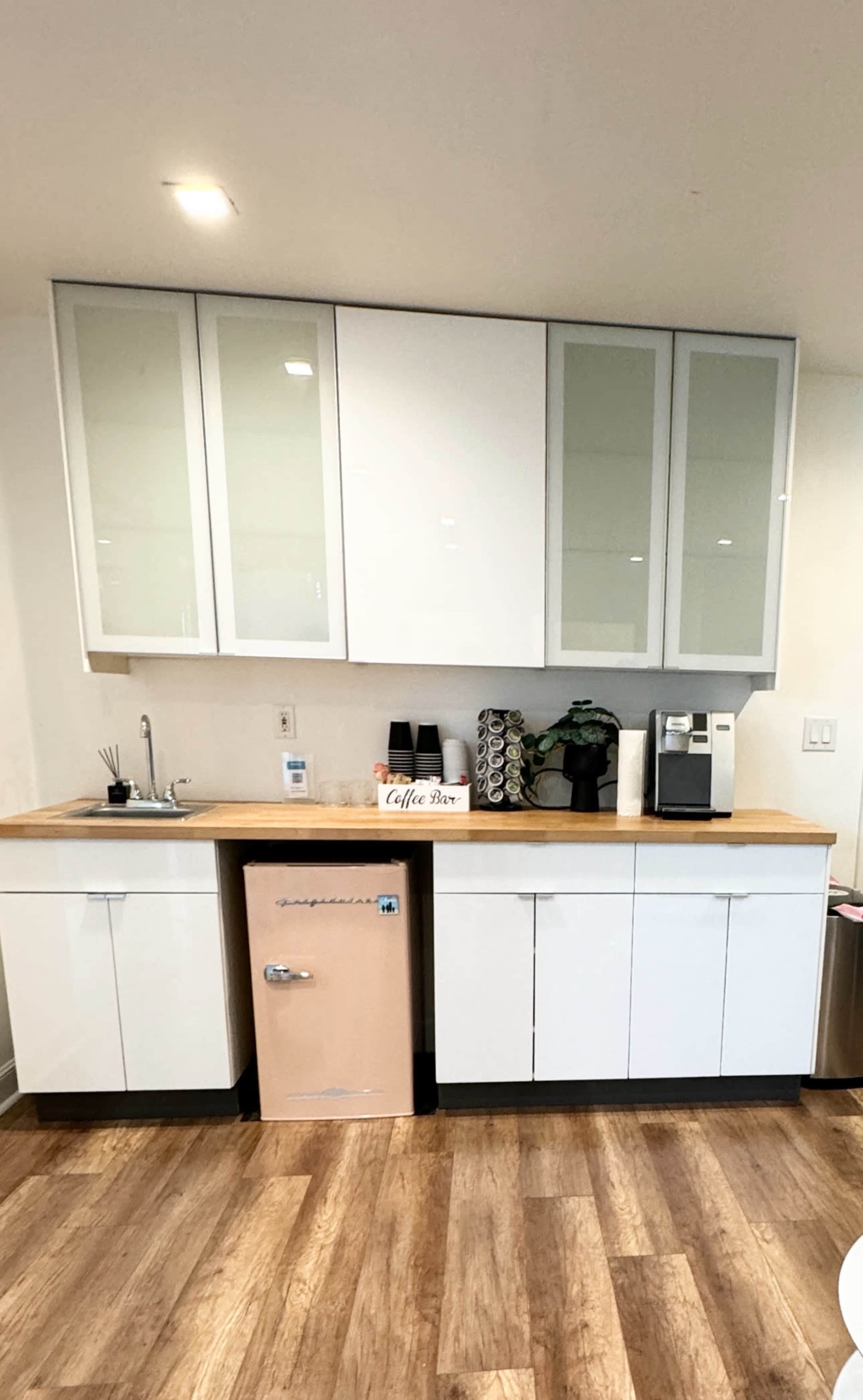 A modern kitchen area with white cabinetry, a wooden countertop, a small sink, a pink refrigerator, and a coffee station.