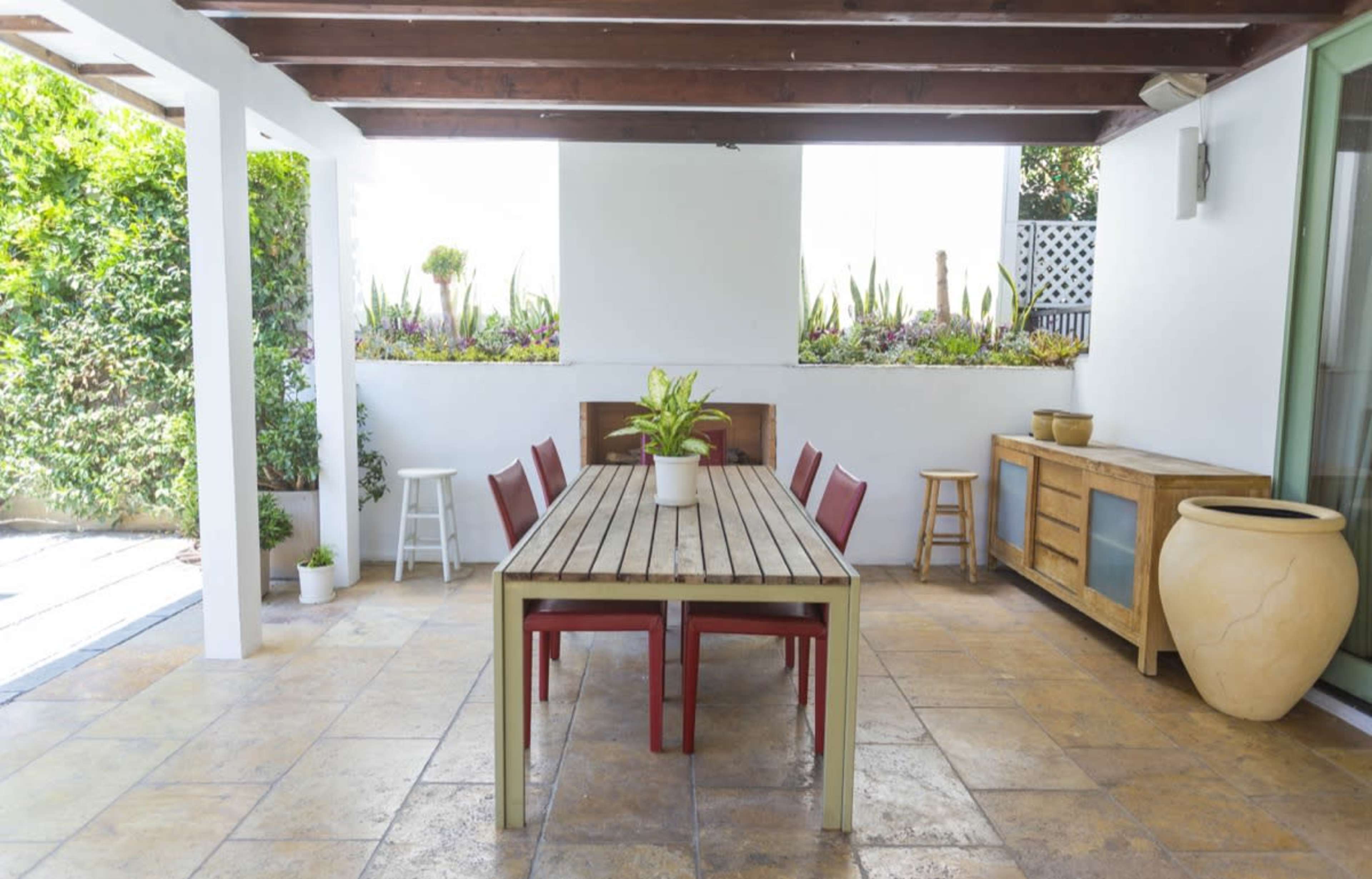 A spacious outdoor dining area features a long wooden table accompanied by red chairs, set under a covered patio with potted plants and a stone floor.