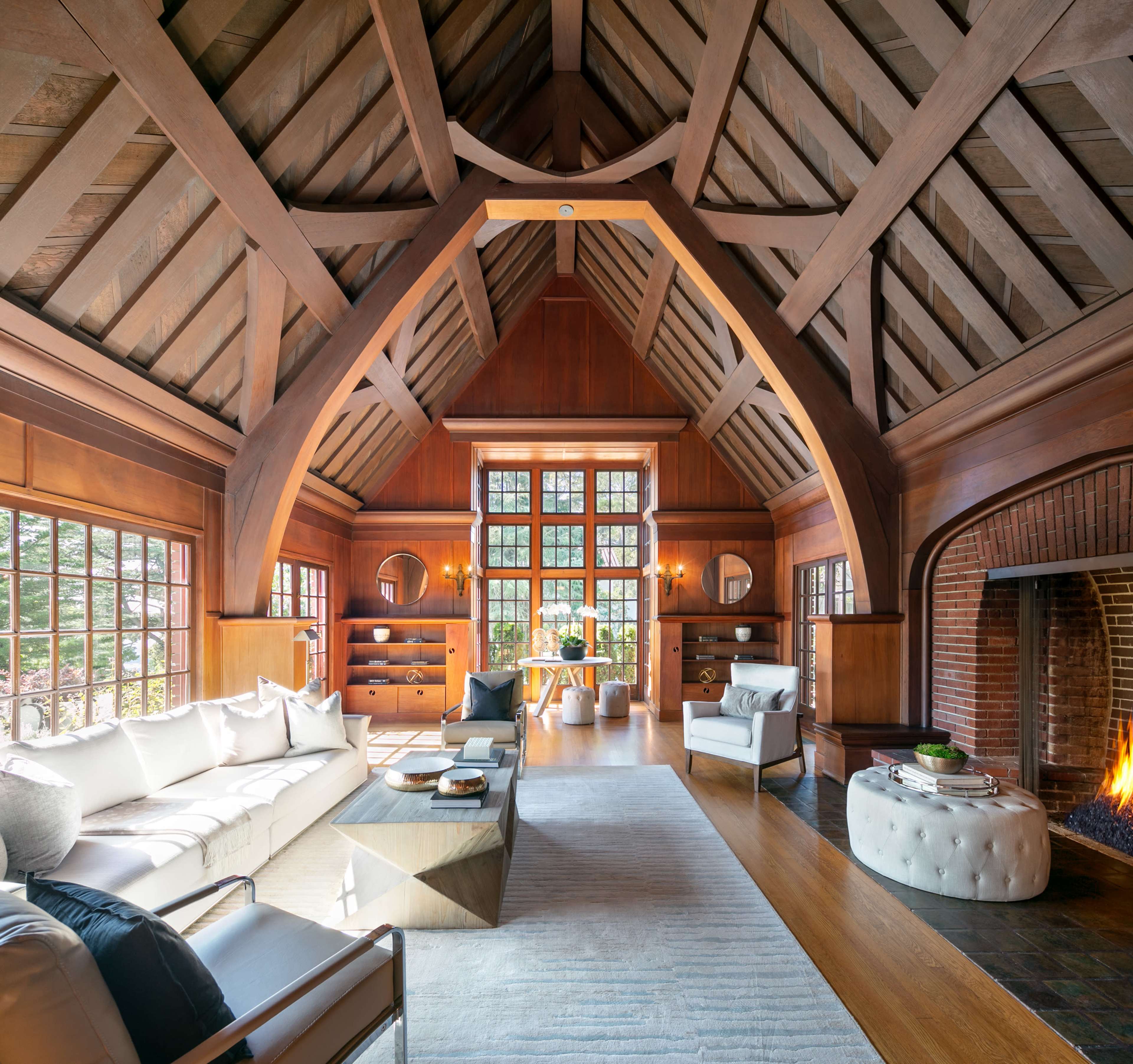 A spacious living room with high wooden-beamed ceilings, large windows, and a fireplace, furnished with modern seating and a coffee table.