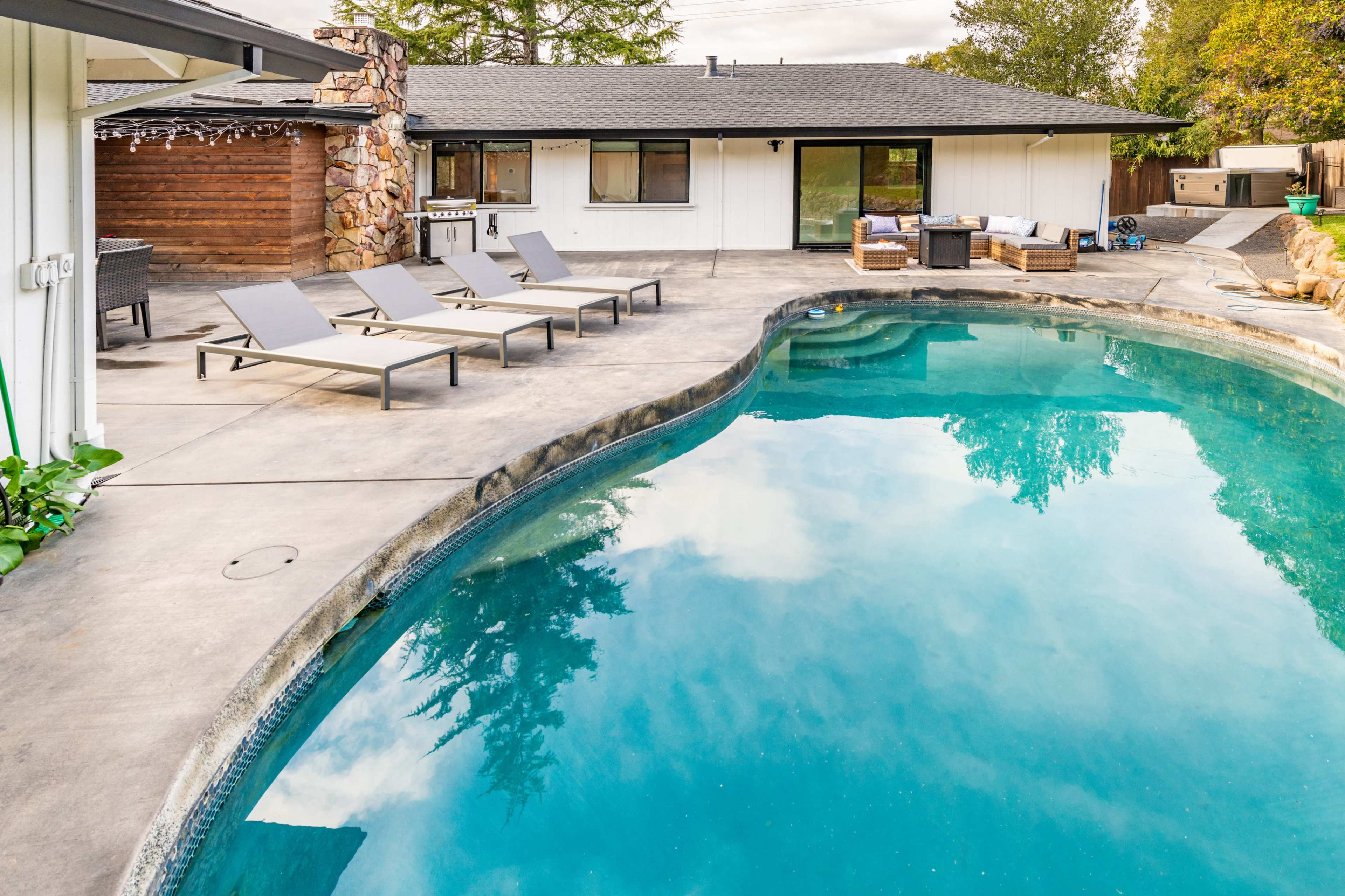 A rectangular swimming pool with lounge chairs surrounds a modern home featuring a stone accent wall and a patio area.