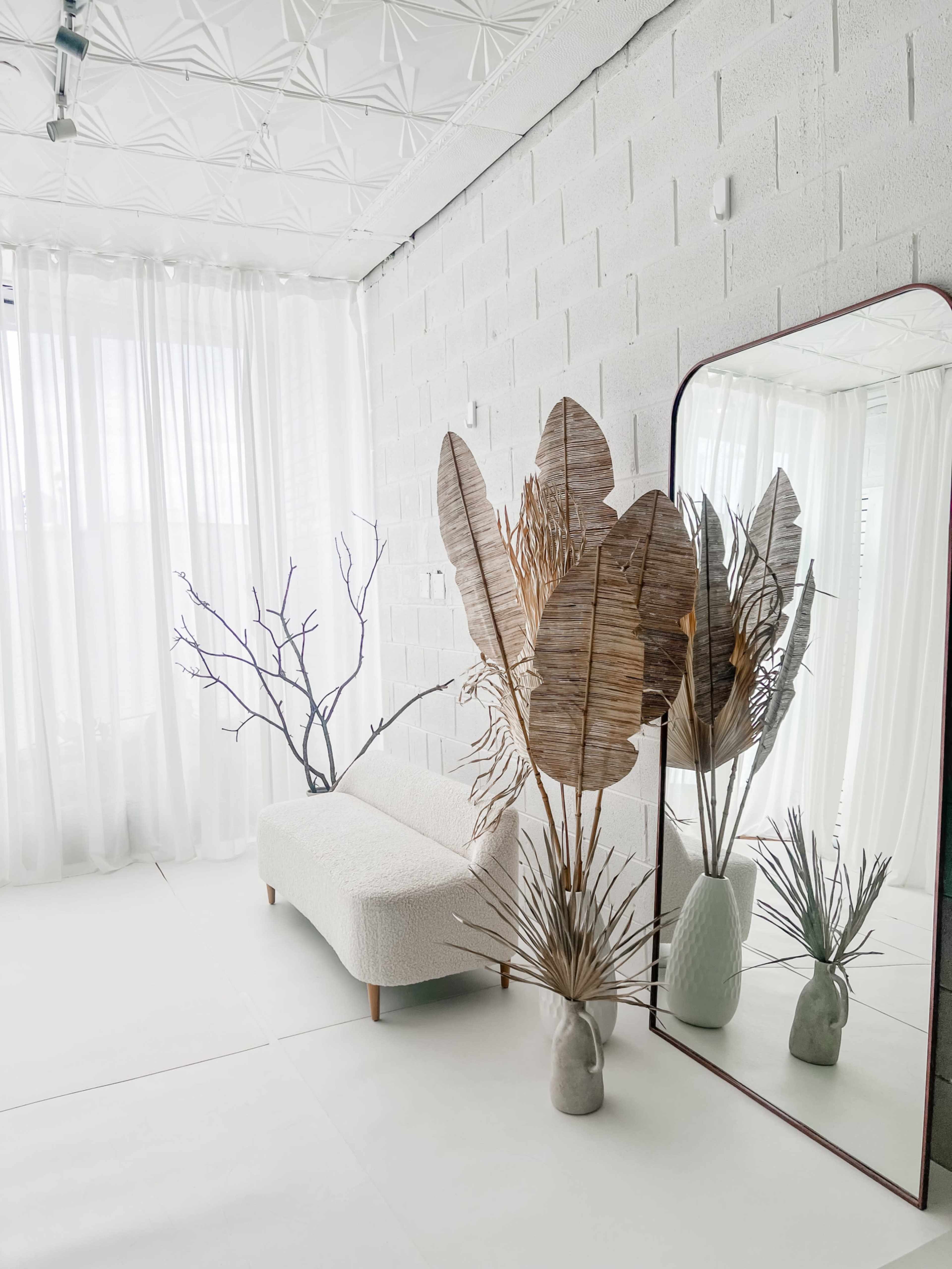 The scene features a minimalist interior with a textured couch, tall dried plants, and a mirror reflecting the decor against a backdrop of sheer white curtains and concrete walls.