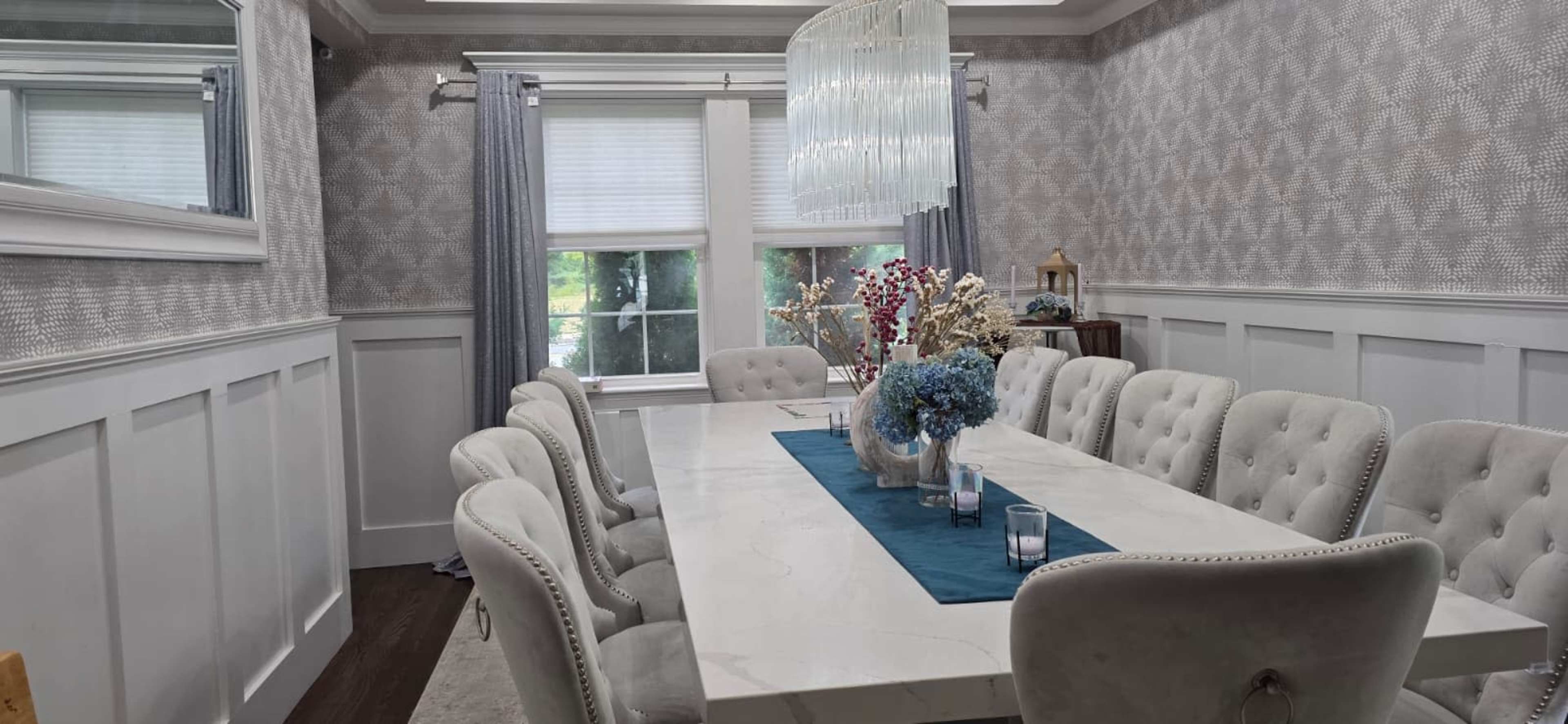 The image shows a dining room with a long, white table surrounded by upholstered chairs, set against patterned wallpaper and a large chandelier overhead.