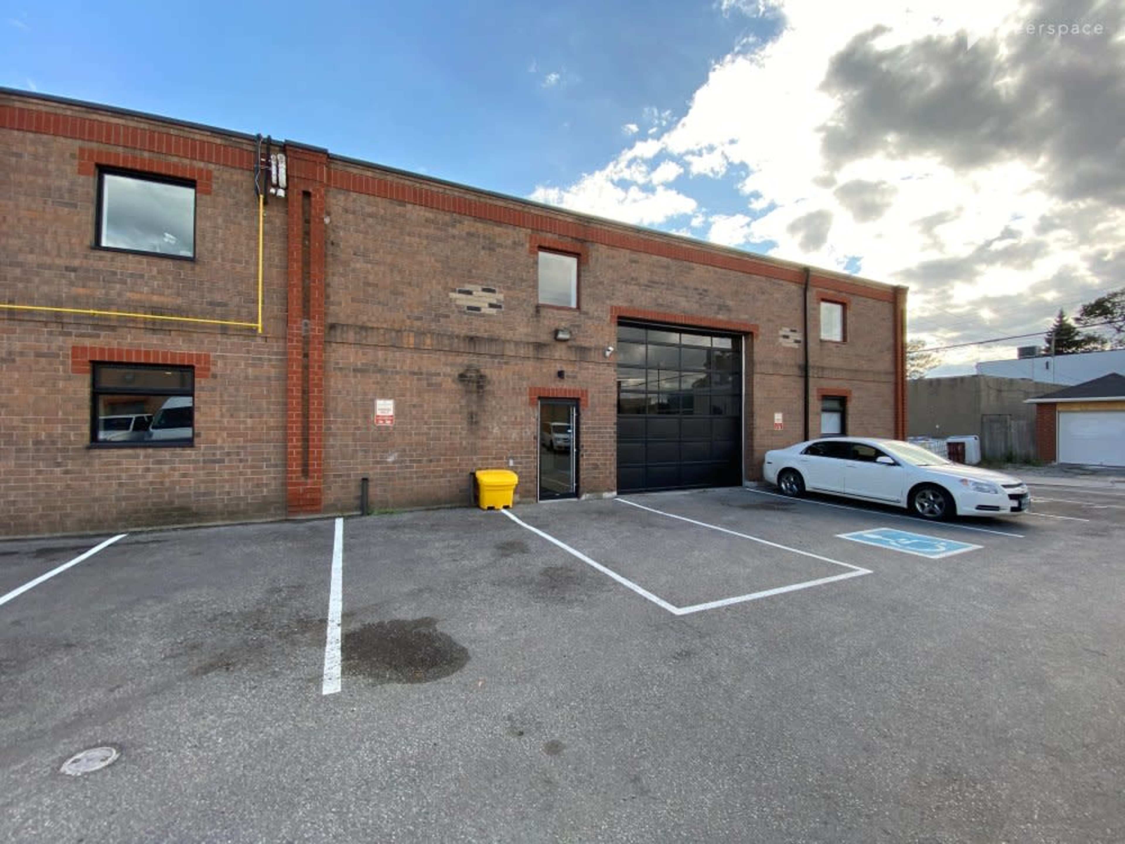 A brick building with a large black garage door, a white car parked in the lot, and a yellow waste bin nearby.