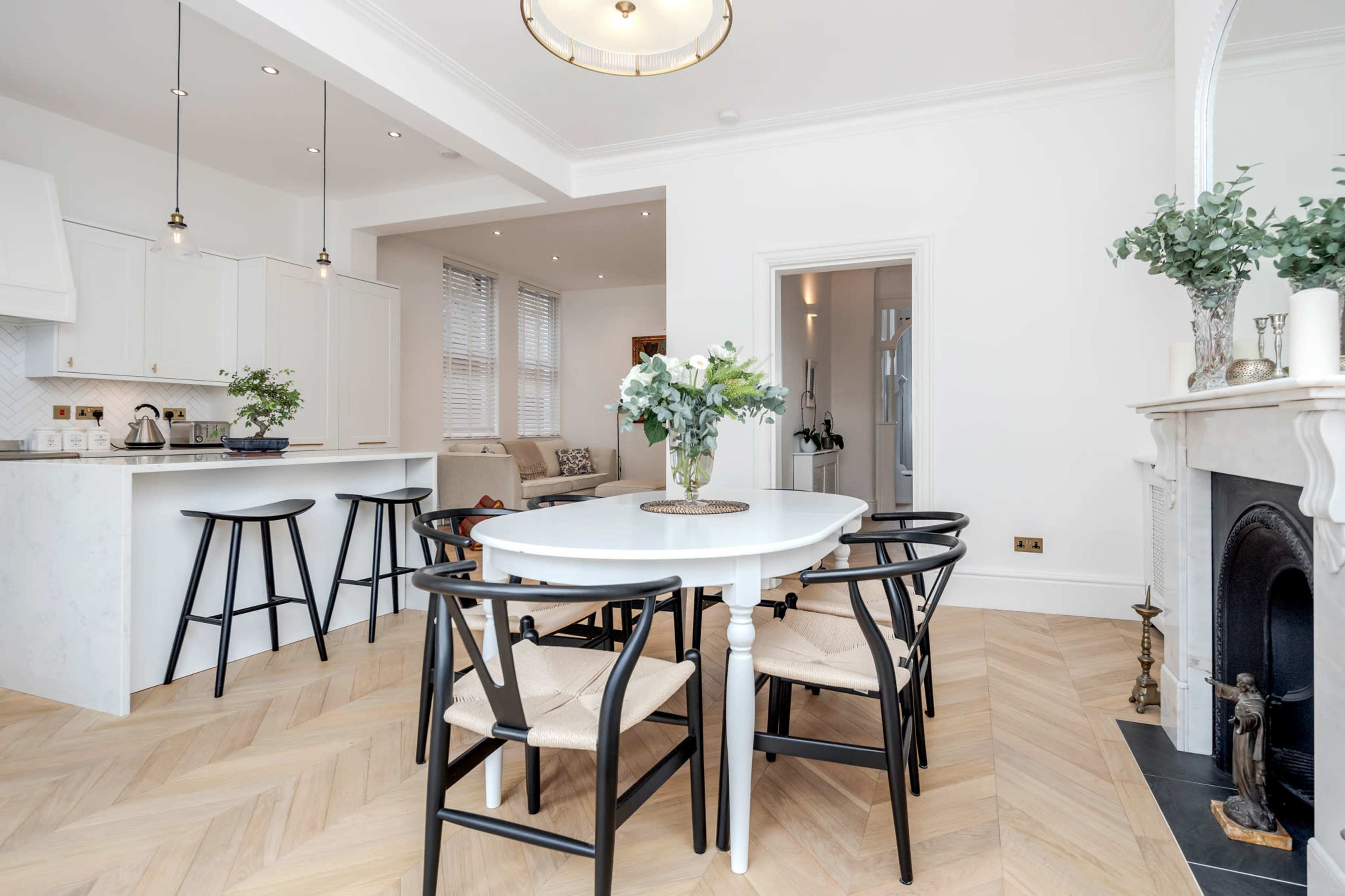 A modern dining area features a round white table surrounded by black chairs, with a light fixture overhead and a cozy living space in the background.