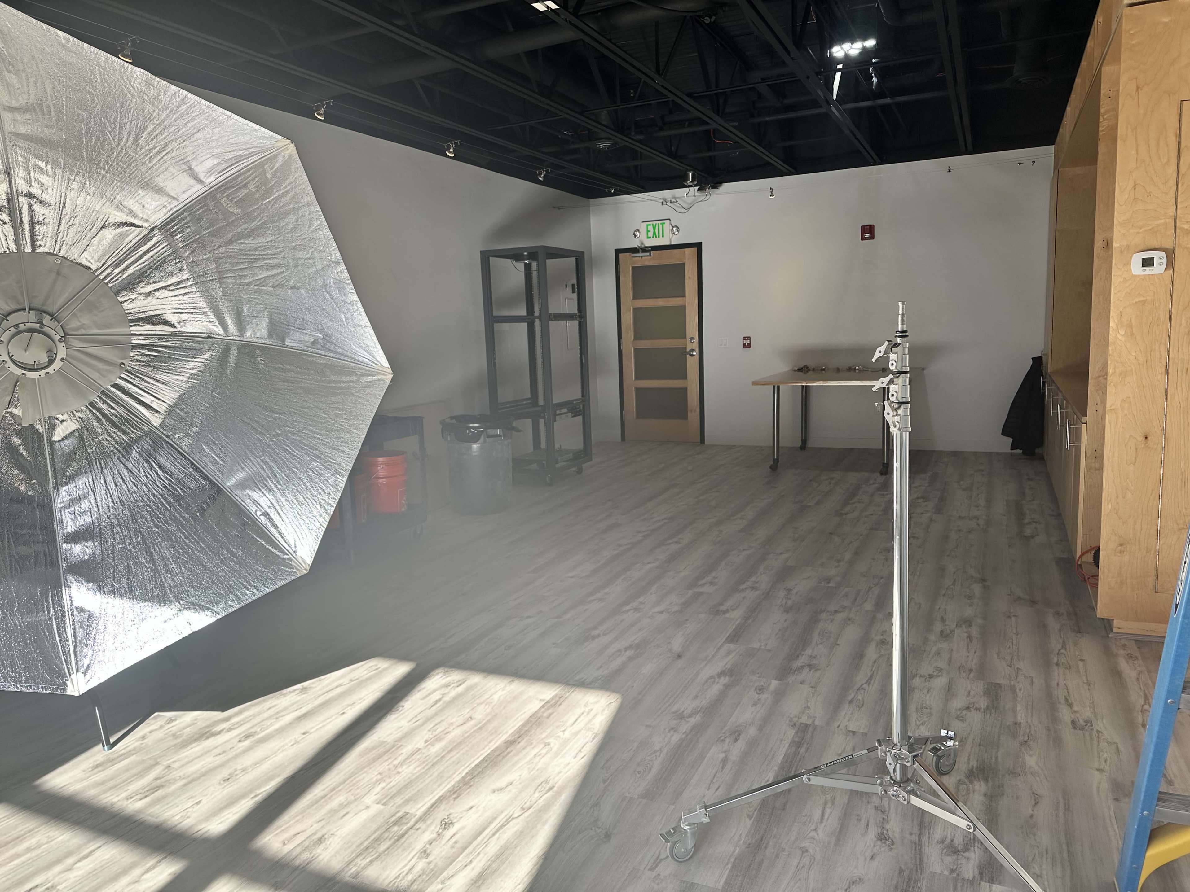 A well-lit studio space with a large reflective umbrella on the left, a light stand, a table, and storage shelves against the wall.