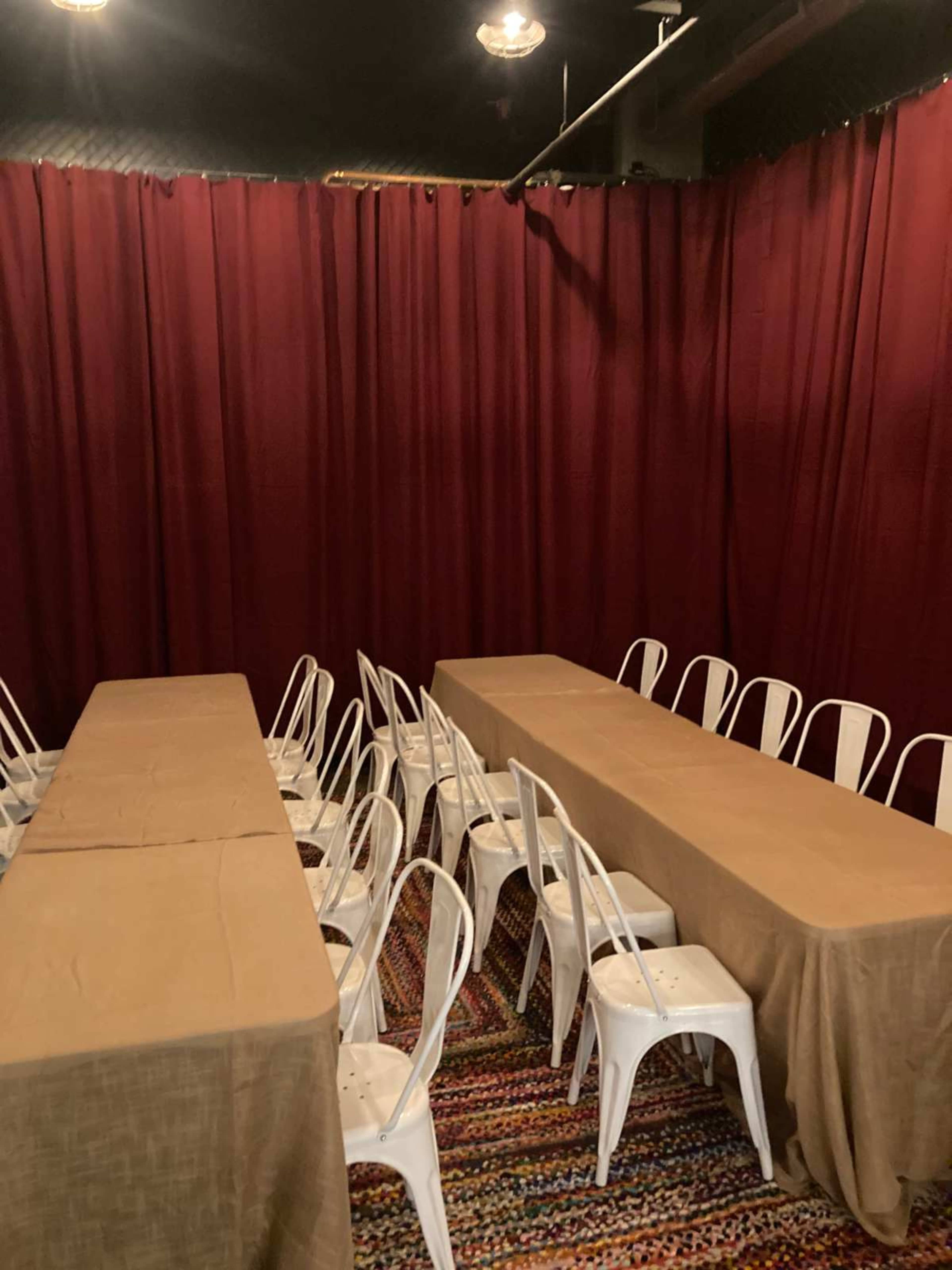 The image shows a room with red curtains, featuring several long tables covered with beige tablecloths and surrounded by white chairs.