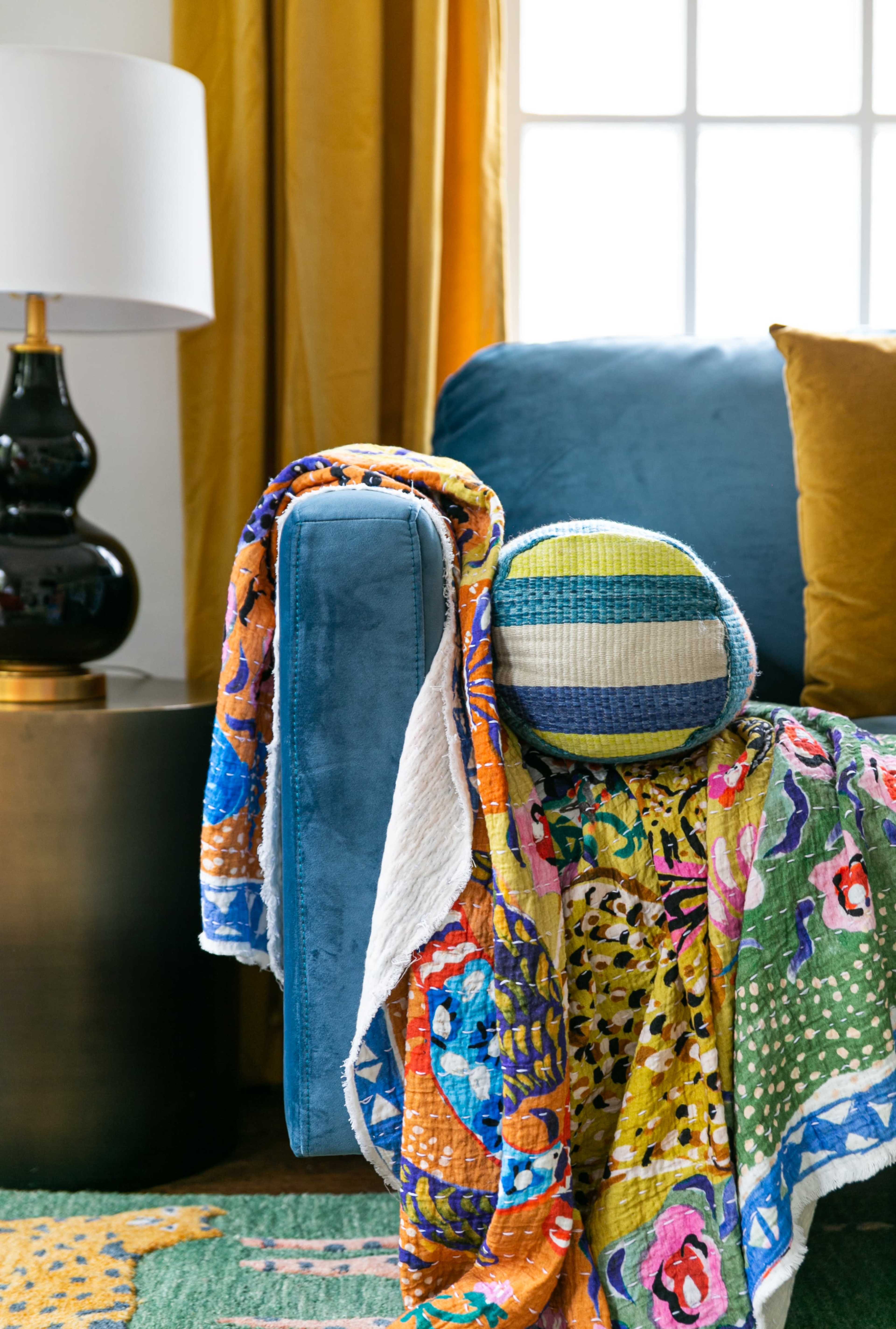 A colorful quilt drapes over a blue couch, with a round woven pillow resting against the arm and a lamp beside it.