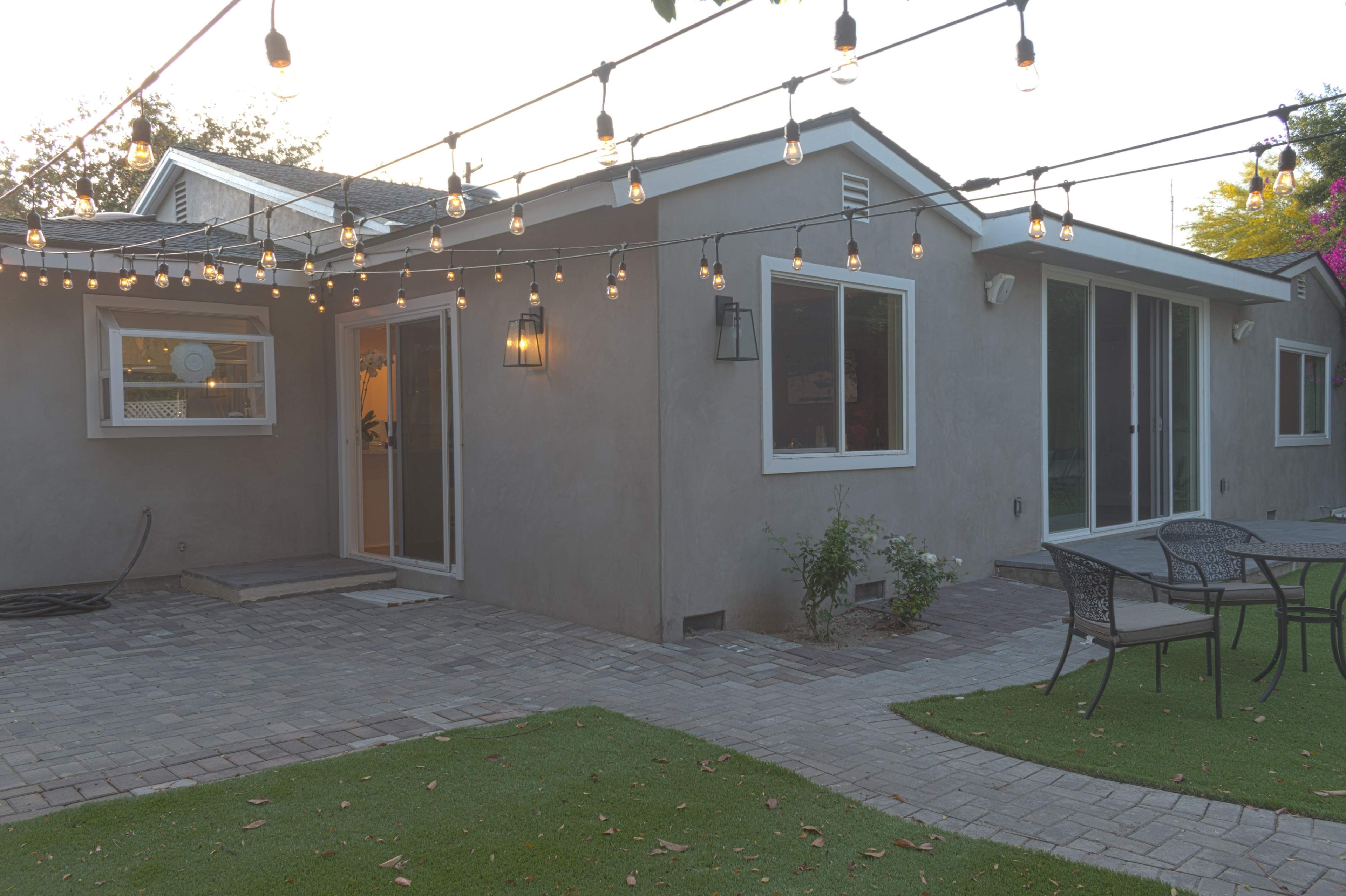 A modern gray house features string lights overhead, with a paved patio area and outdoor seating.