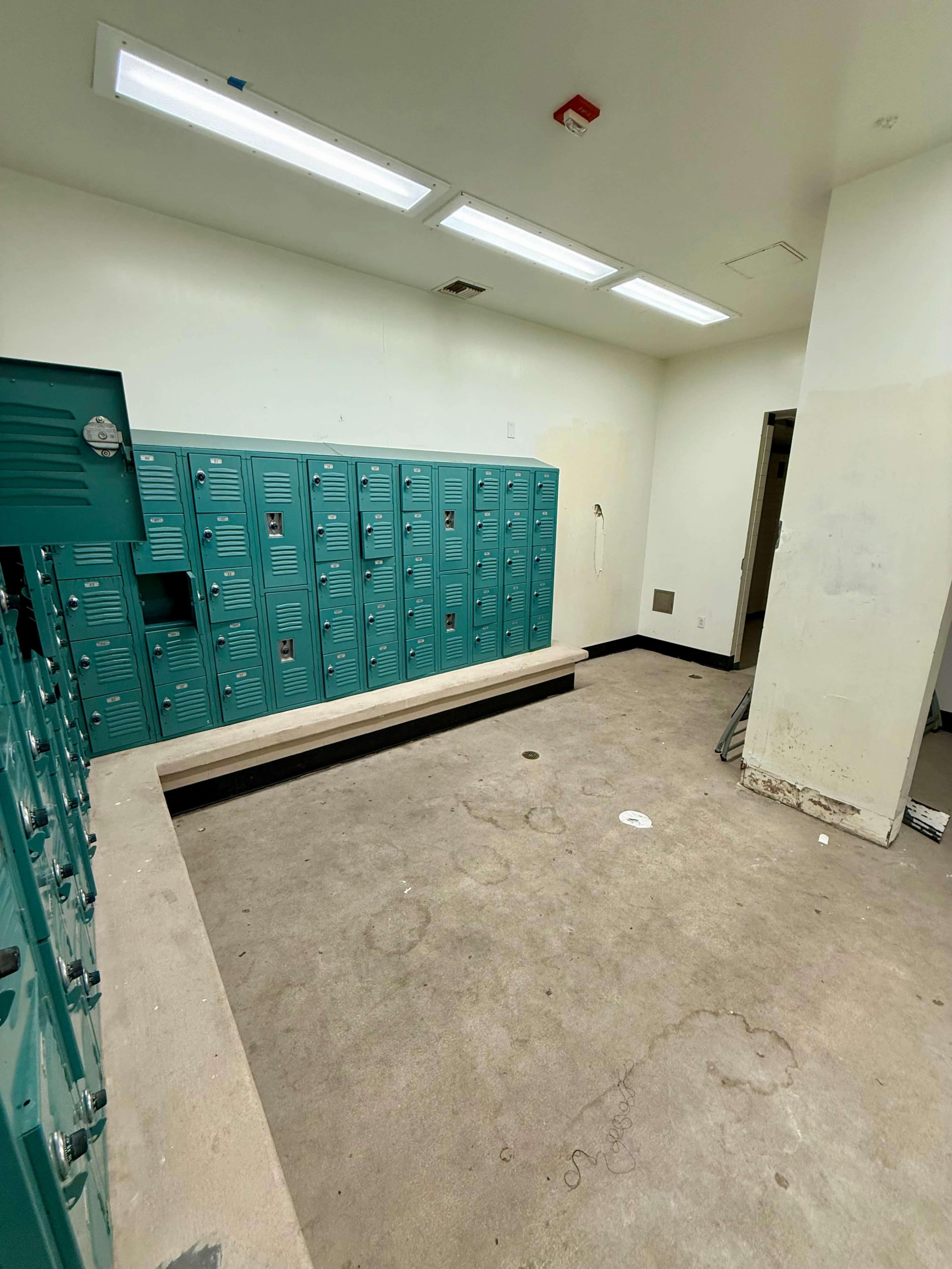 The image shows a room with teal lockers lining one wall and a concrete bench against the opposite wall.