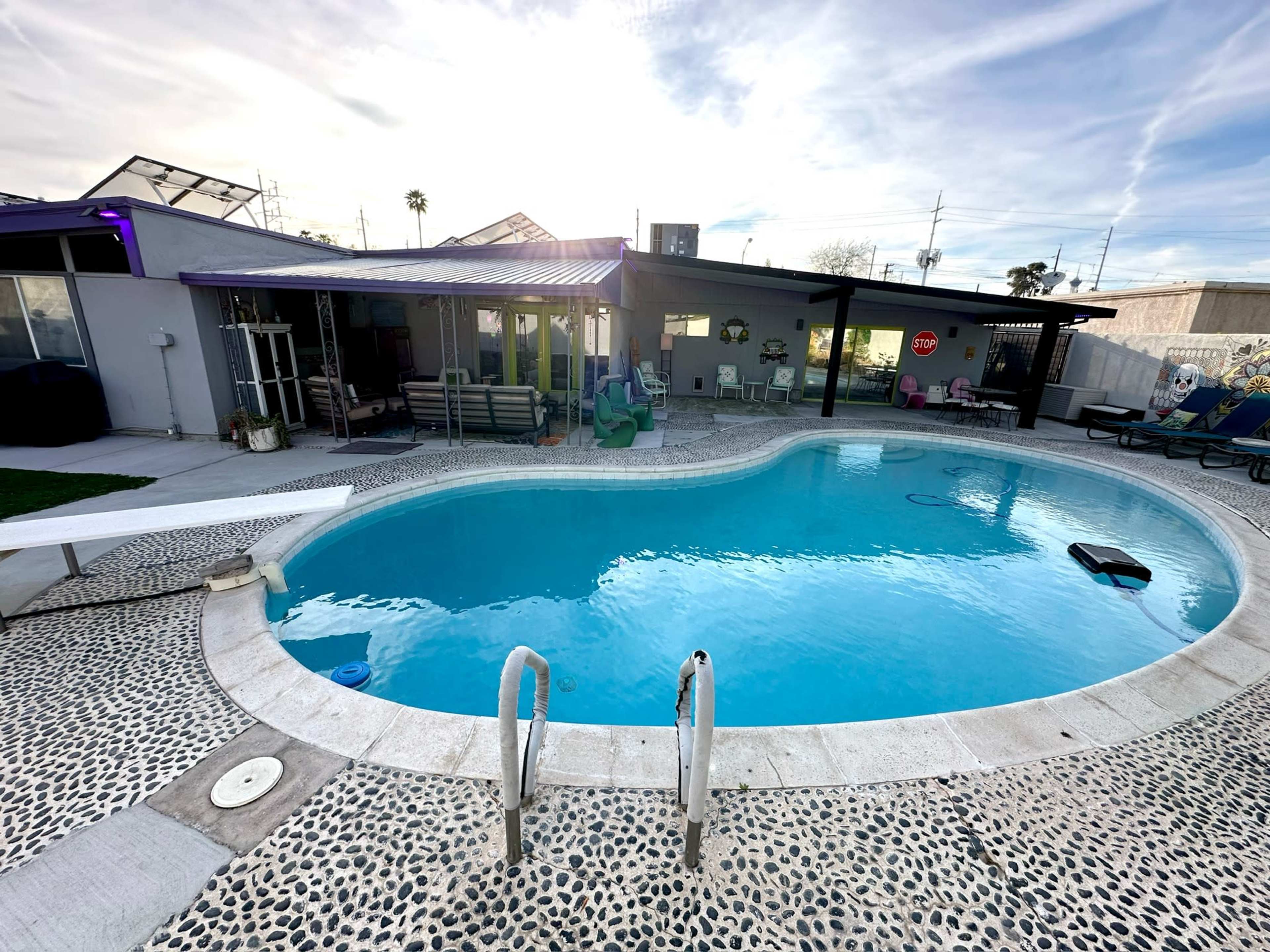 A swimming pool with a diving board and a surrounding patio area is situated beside a covered seating area and lounge chairs.