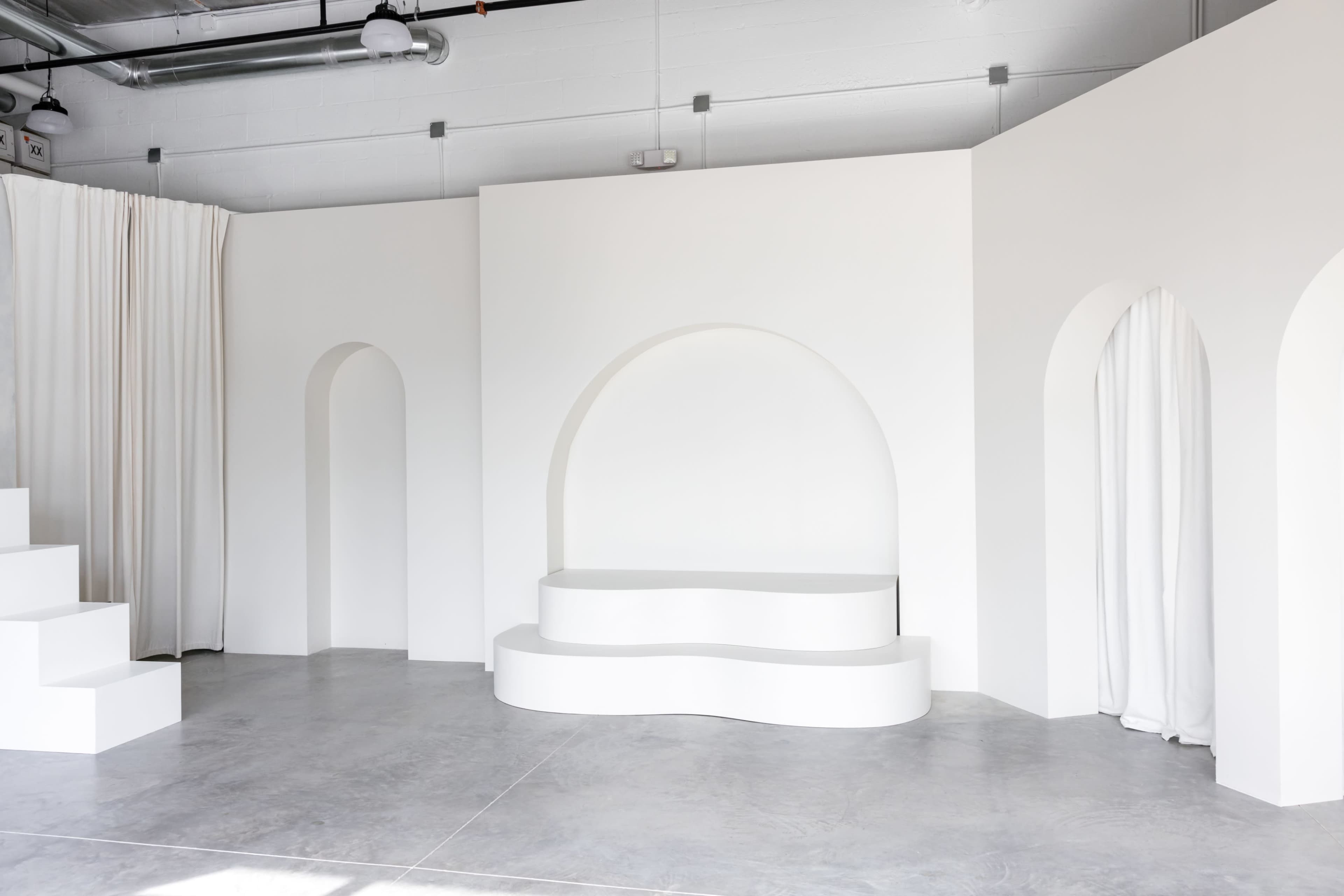 A minimalist, white architectural interior featuring curved and angular walls, an arched niche, and a stepped platform.