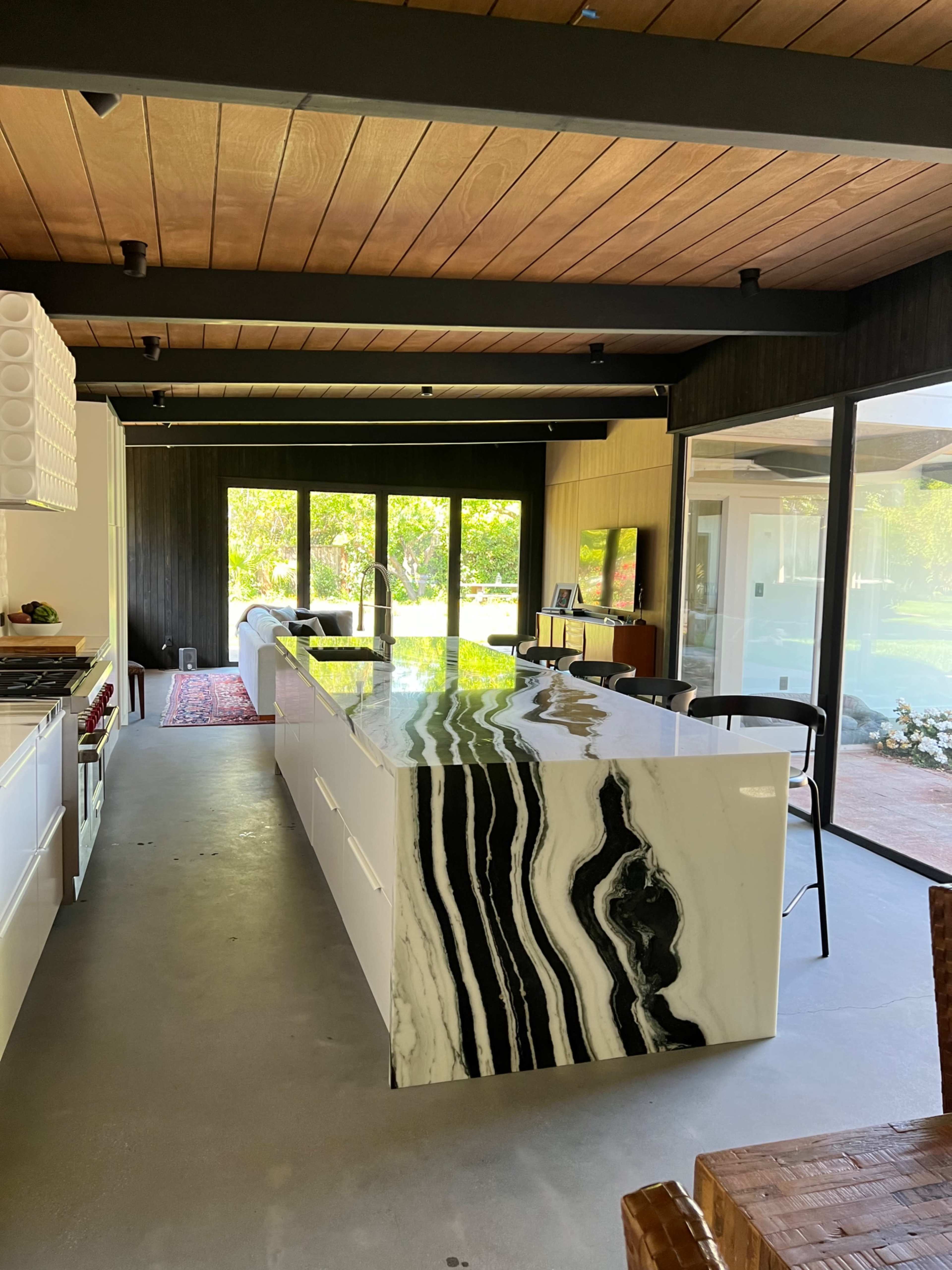 A modern kitchen featuring a black and white marble island, large windows overlooking a garden, and sleek cabinetry.