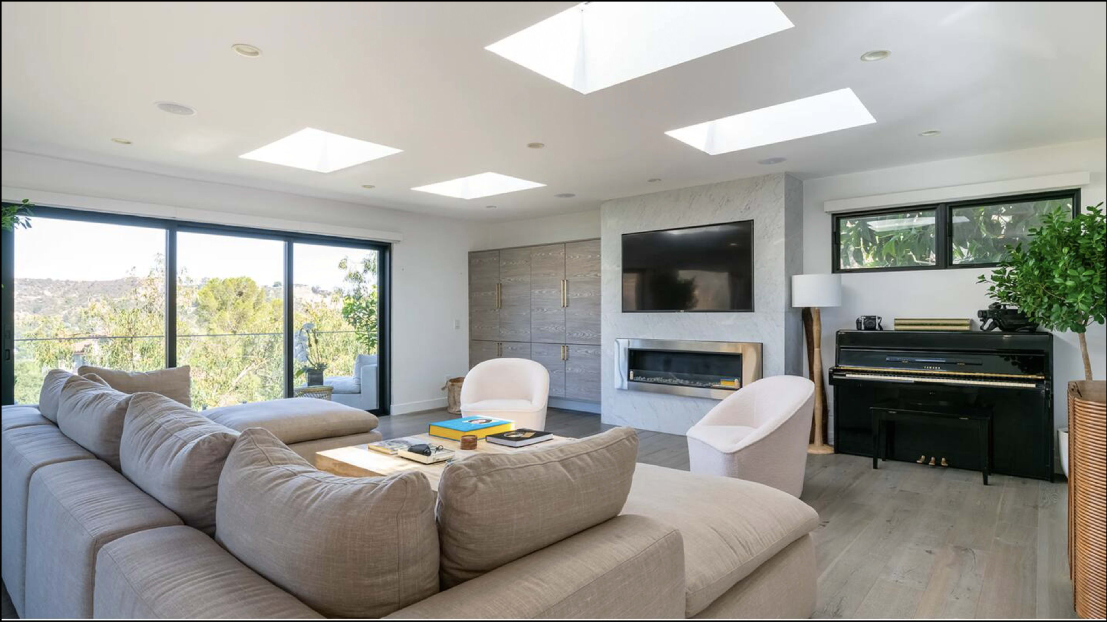 The living room features a large sectional sofa, a modern fireplace, a piano, and natural light coming through skylights and large windows.