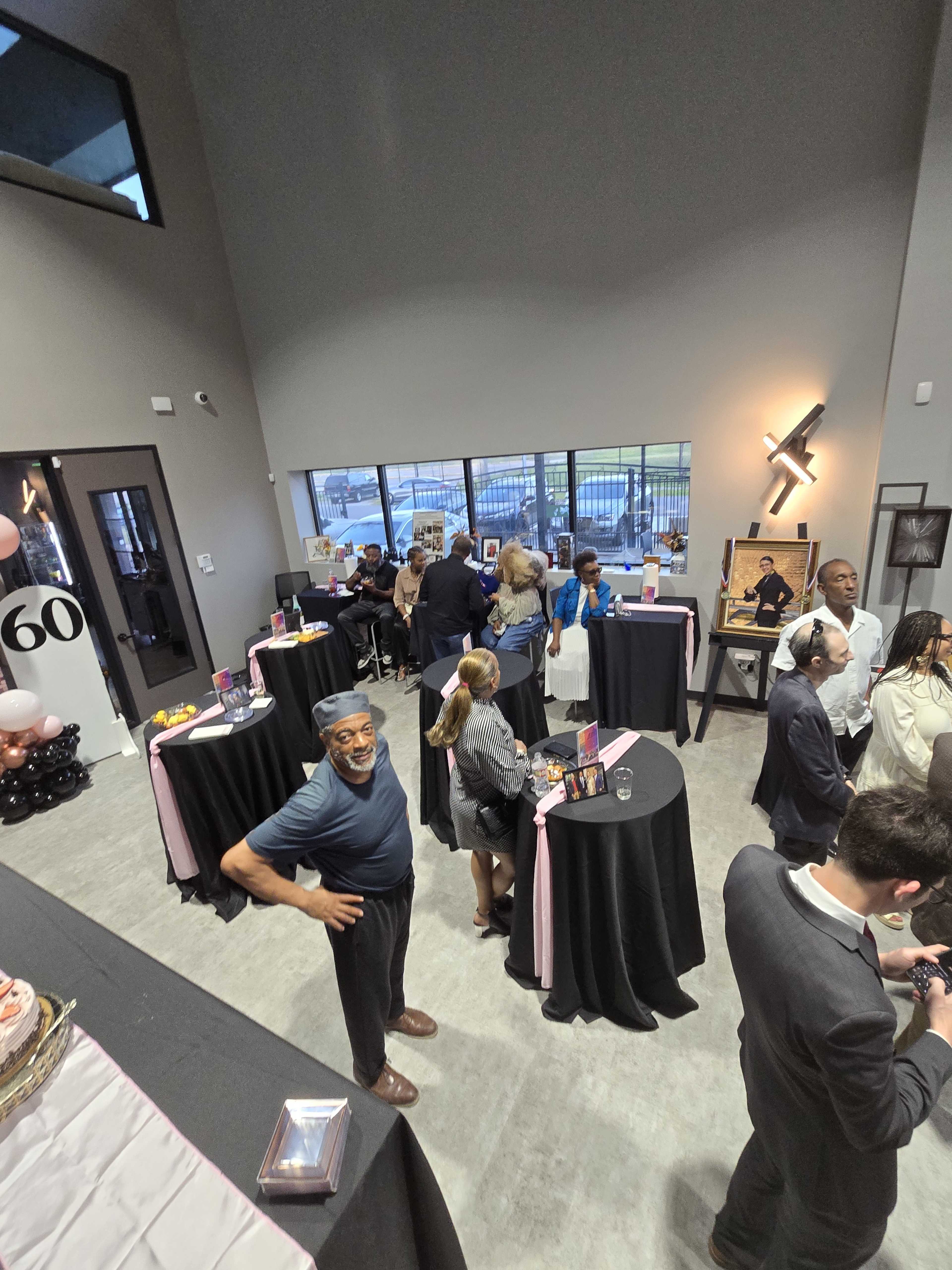 The image shows a gathering of people mingling around black tables in a modern event space with large windows.