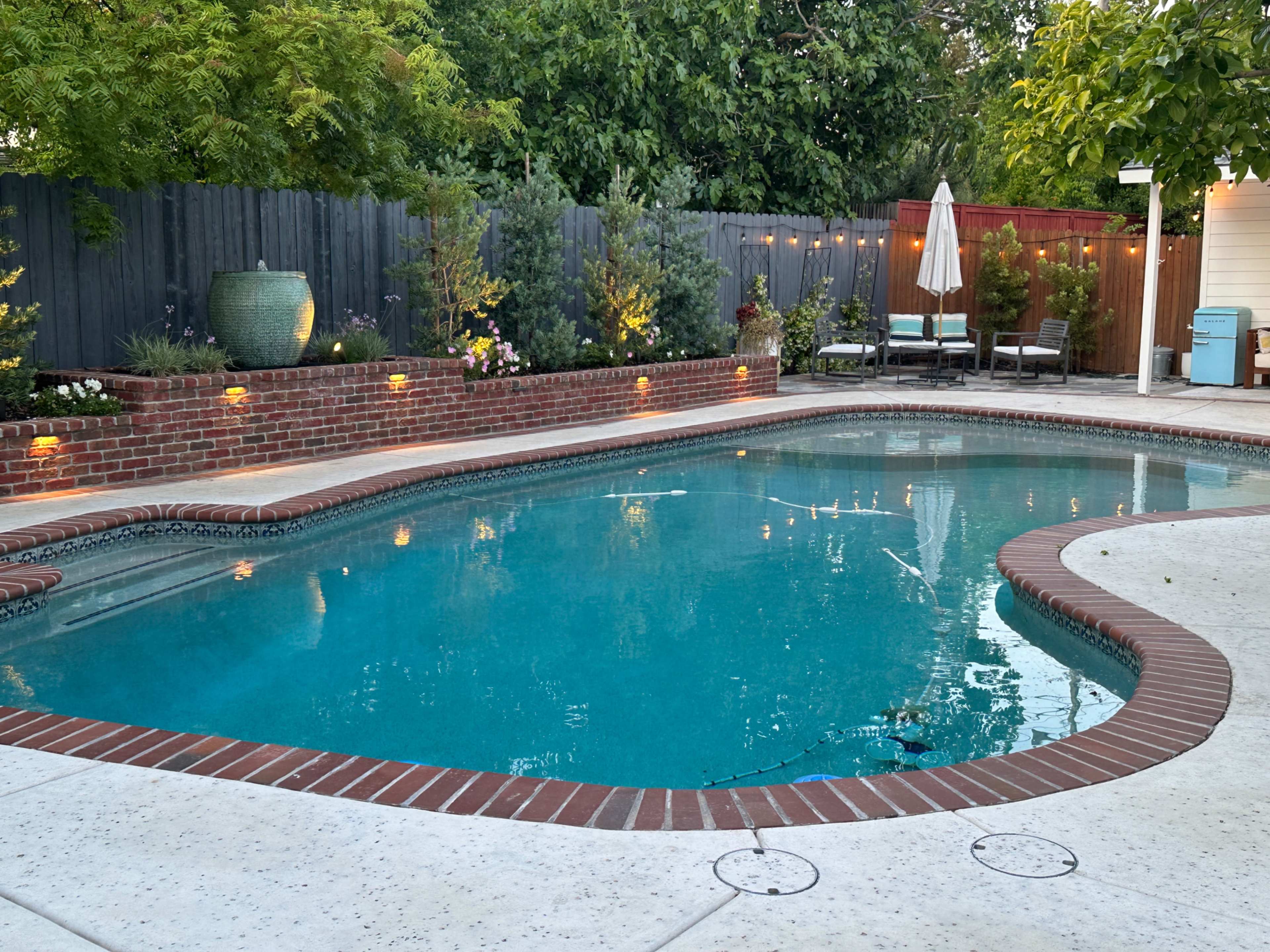 The image shows a residential swimming pool surrounded by a landscaped patio area with garden lights and lounge chairs.